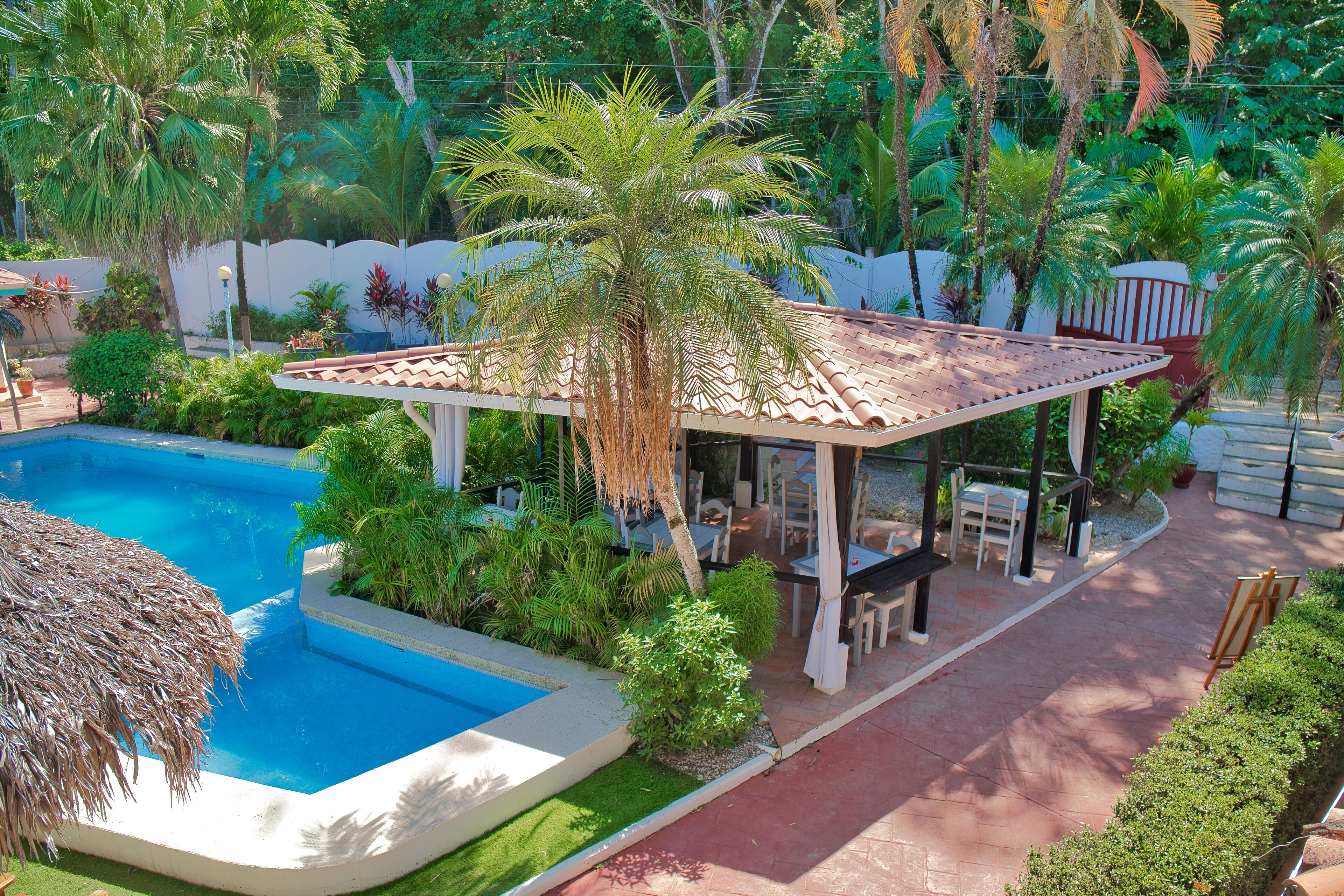 A pool area with a covered seating area, surrounded by lush greenery and palm trees.