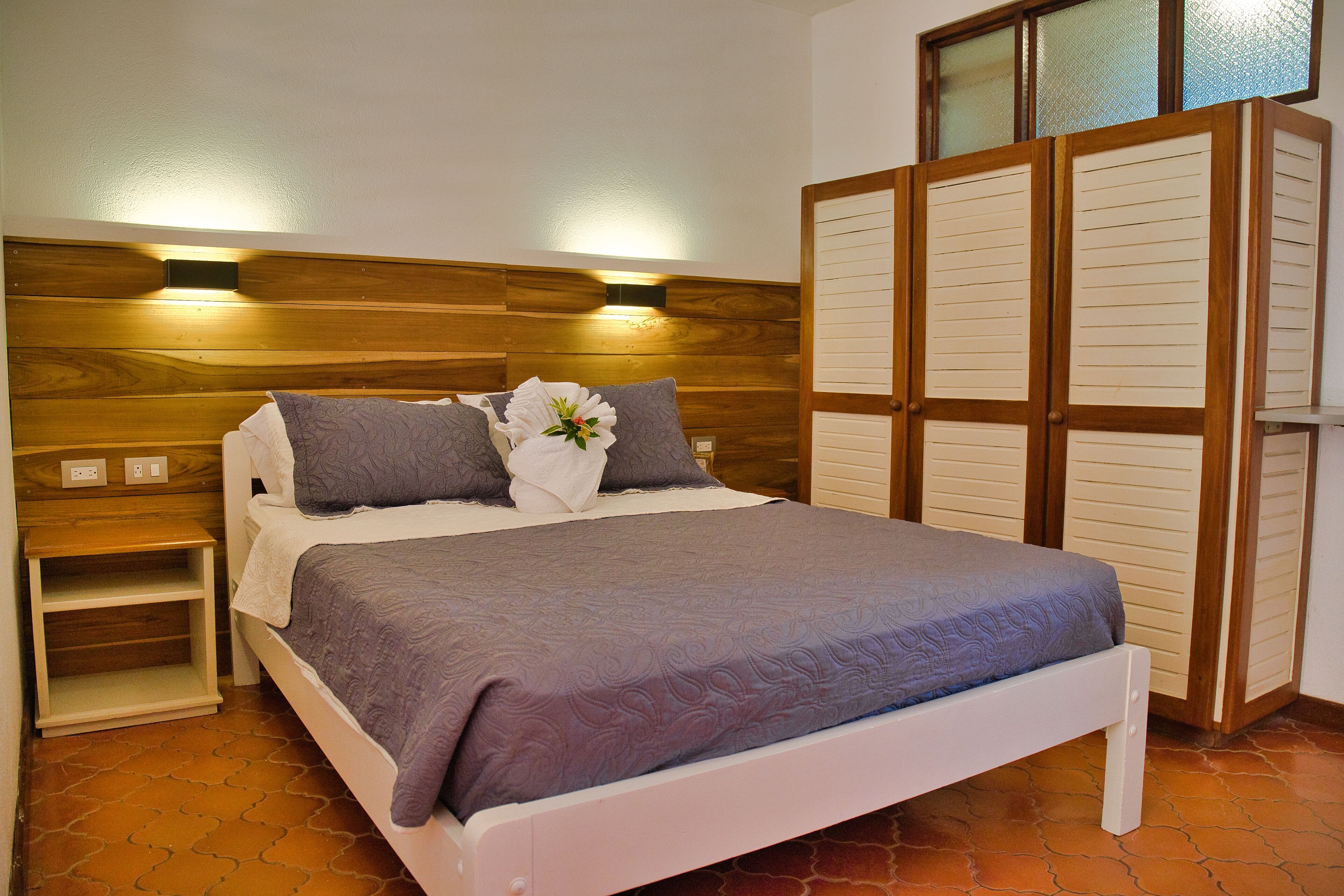 Ein Schlafzimmer mit einem hölzernen Kopfteil, einem Bett mit grauer Bettdecke, einem Nachttisch und einem Schrank mit Klappläden.