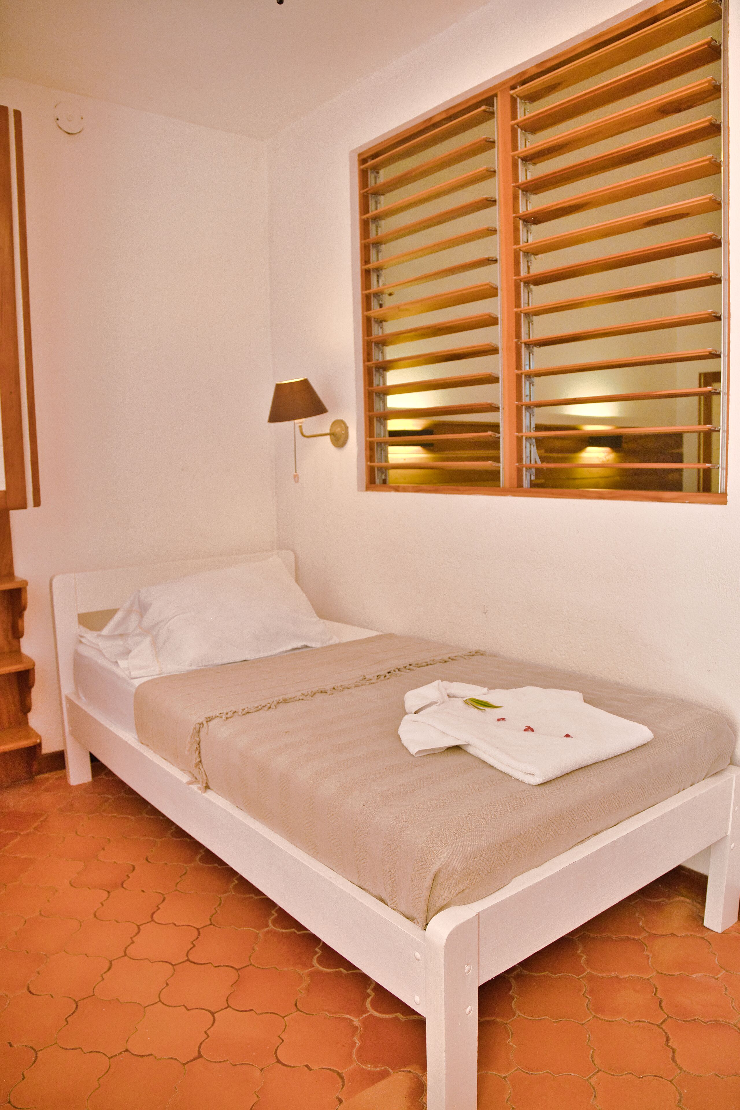 A single-bed room with a wooden headboard, a bedside lamp, and a window with wooden blinds.