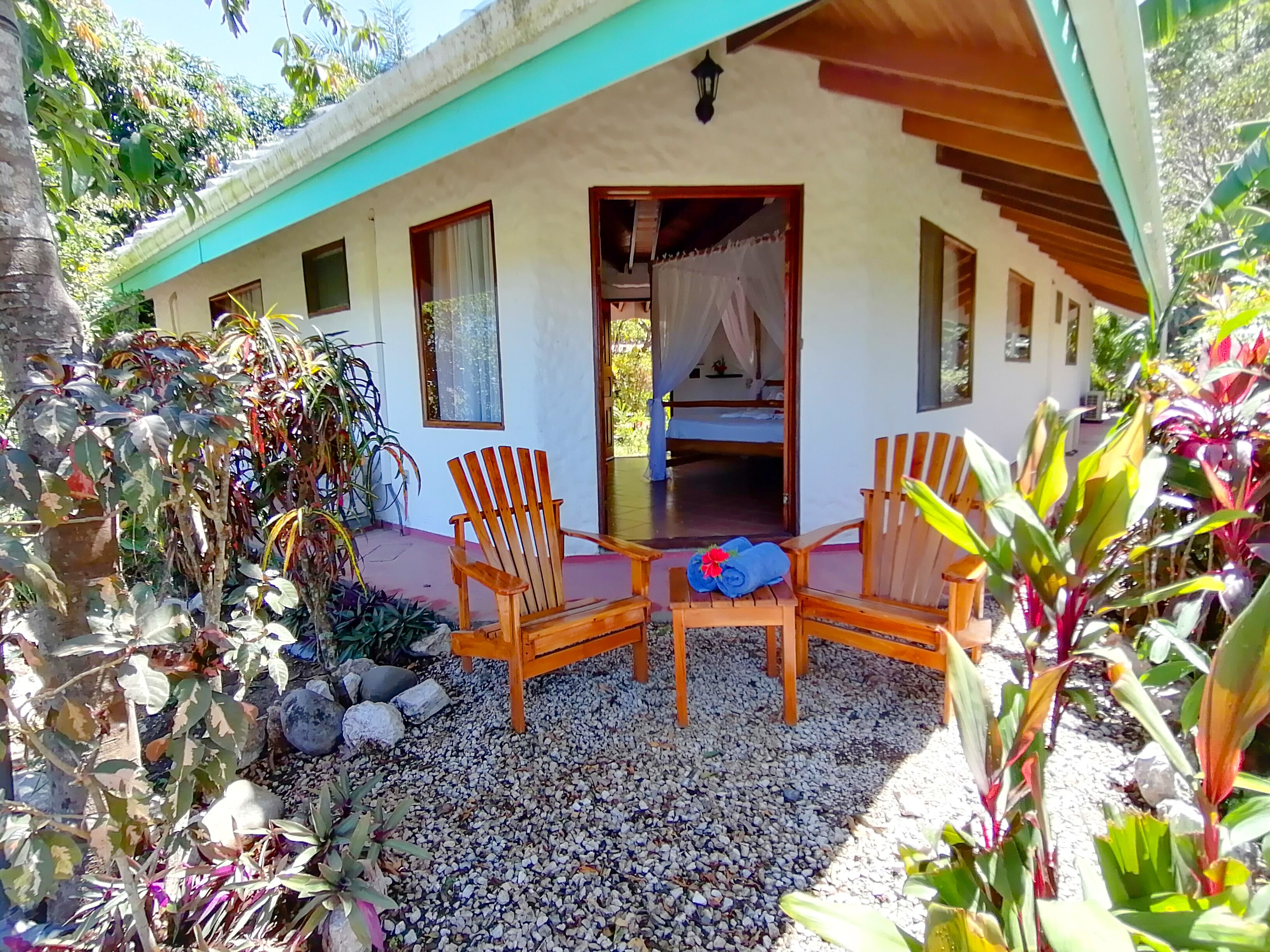 Un bungalow doté d’un porche couvert, de deux chaises en bois, d’une petite table et d’un lit à baldaquin.