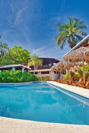 A swimming pool with lounge chairs, palm trees, and a building with a thatched roof. - Hotel Selva y Mar Sámara (Samara)