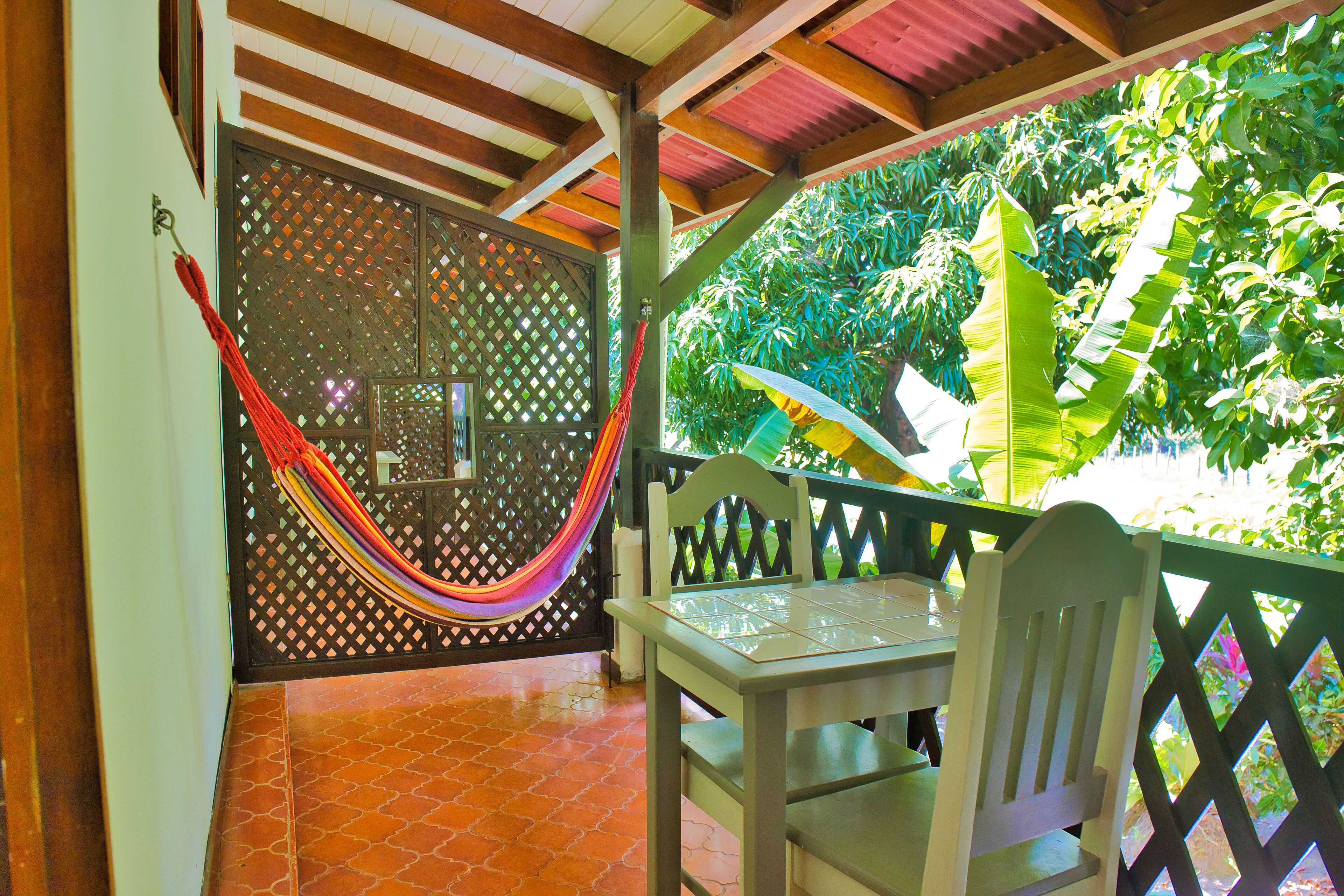 A covered patio with a wooden table and chairs, a hammock, and a lattice door.