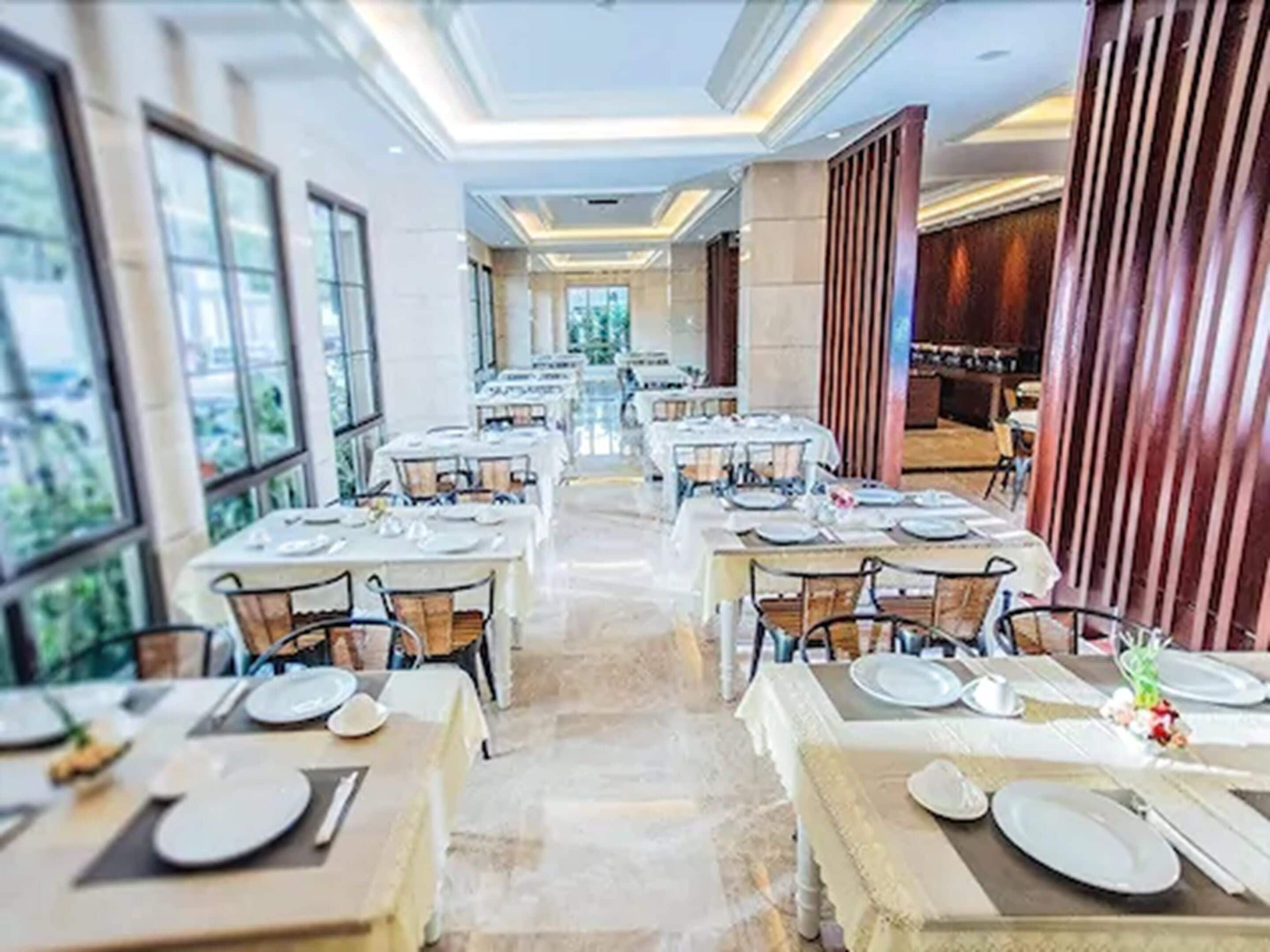 Un restaurant avec de nombreuses tables dressées pour le service, de grandes fenêtres et une vue sur la verdure environnante.