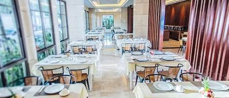 Un restaurant avec de nombreuses tables dressées pour le service, de grandes fenêtres et une vue sur la verdure environnante.