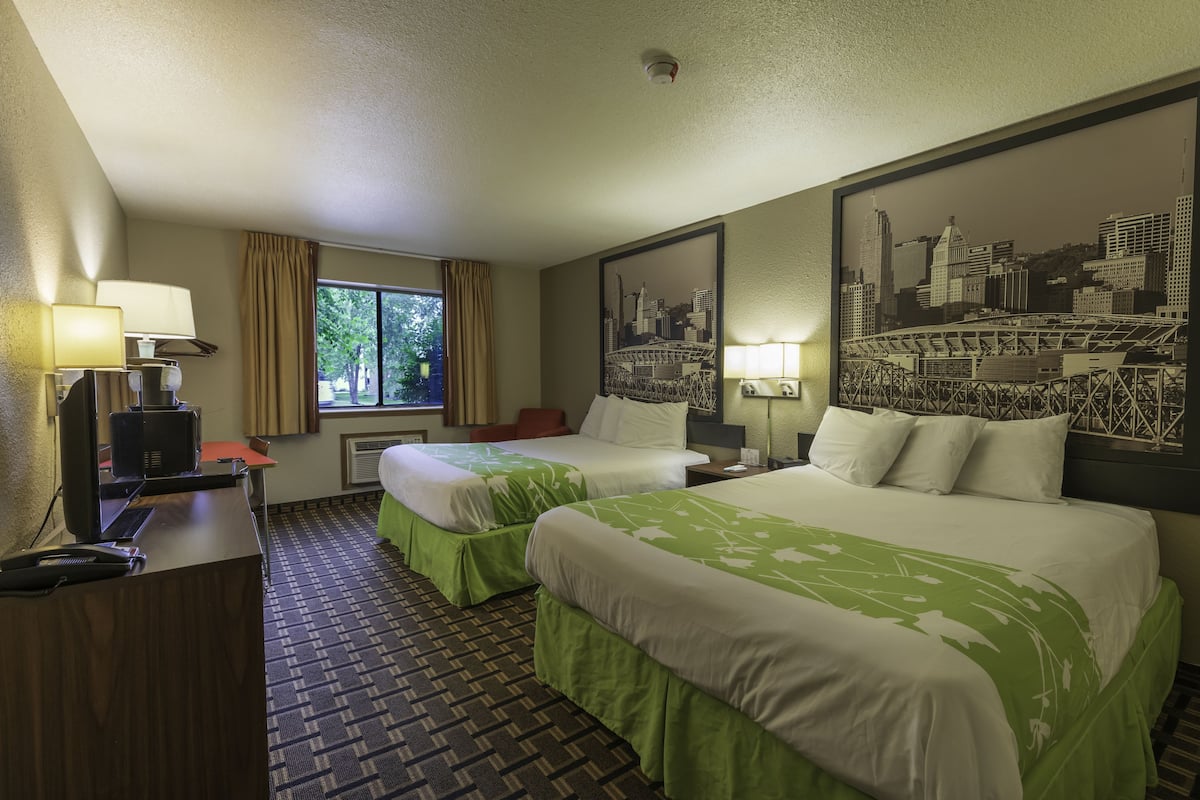 A hotel room with two beds, a desk, a chair, a window with curtains, and a cityscape painting on the wall.