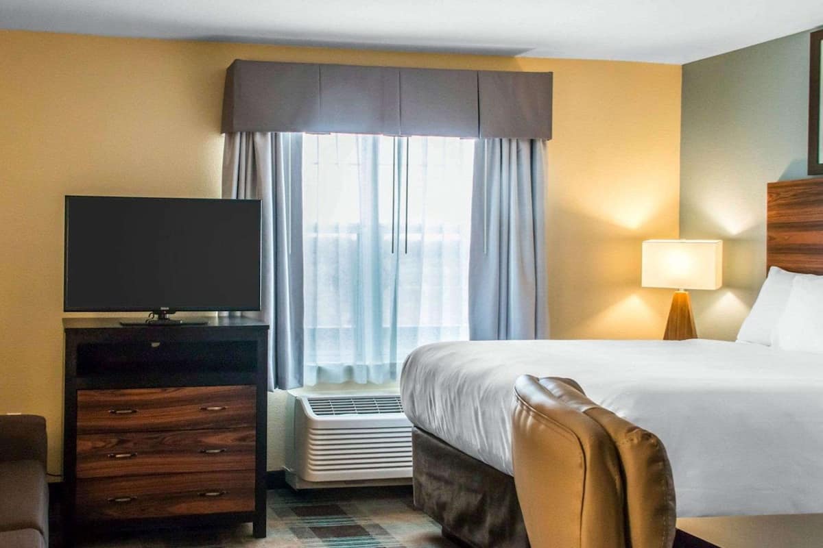 Ein Hotelzimmer mit einem Bett, einem Fernseher, einem Schreibtisch mit Schubladen, einem Sessel, einem Fenster mit Vorhängen und einer Wandlampe.