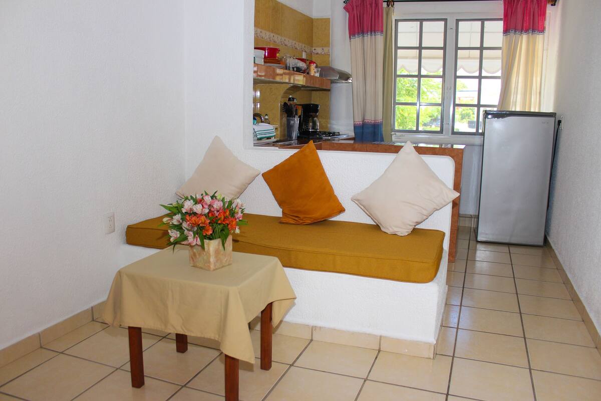 Un espace de vie compact comprenant un canapé, une petite table avec un vase de fleurs, un coin cuisine avec un réfrigérateur et une fenêtre avec des rideaux.