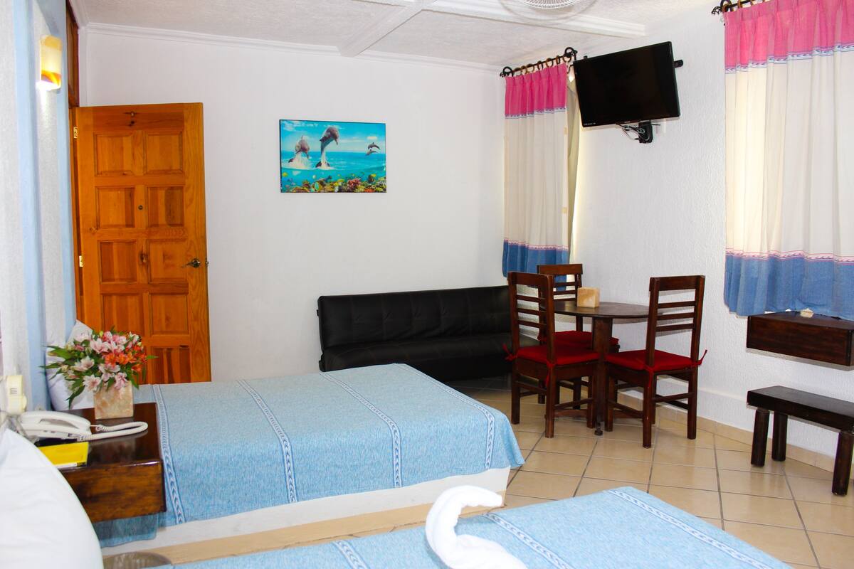 Une chambre d’hôtel avec deux lits, une porte en bois, un tableau, une télévision, une table à manger avec des chaises et un banc.