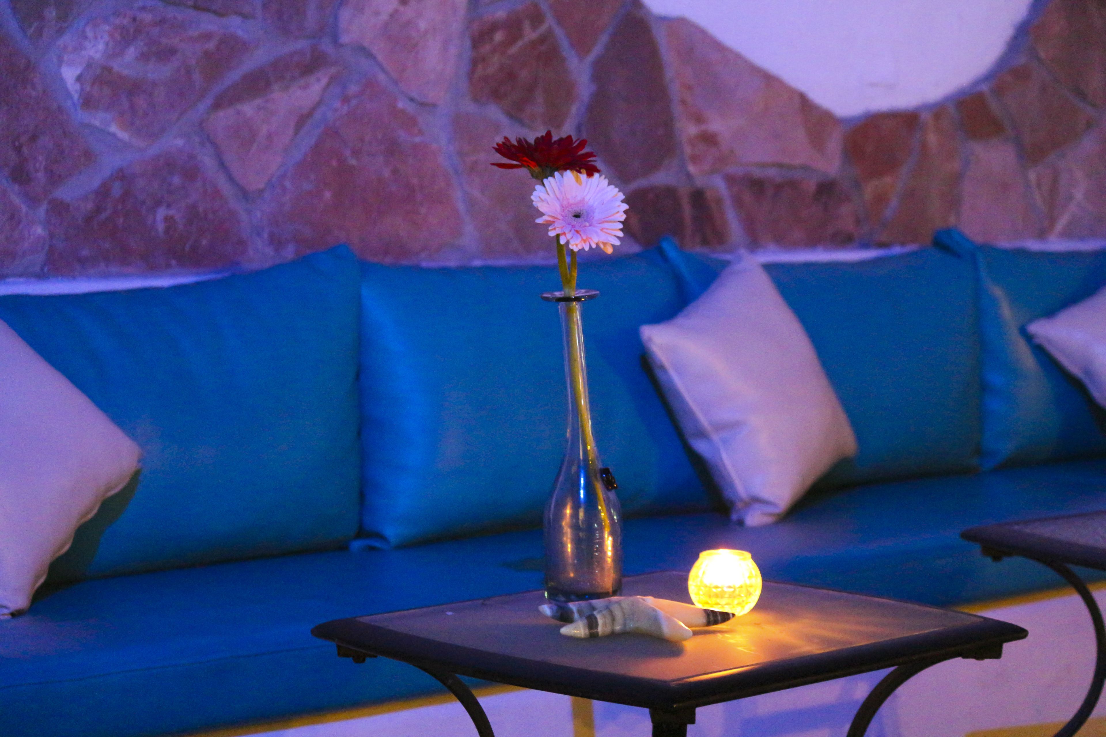 A table with a vase containing flowers and a lit candle, placed in front of a blue sofa with stone wall backdrop.