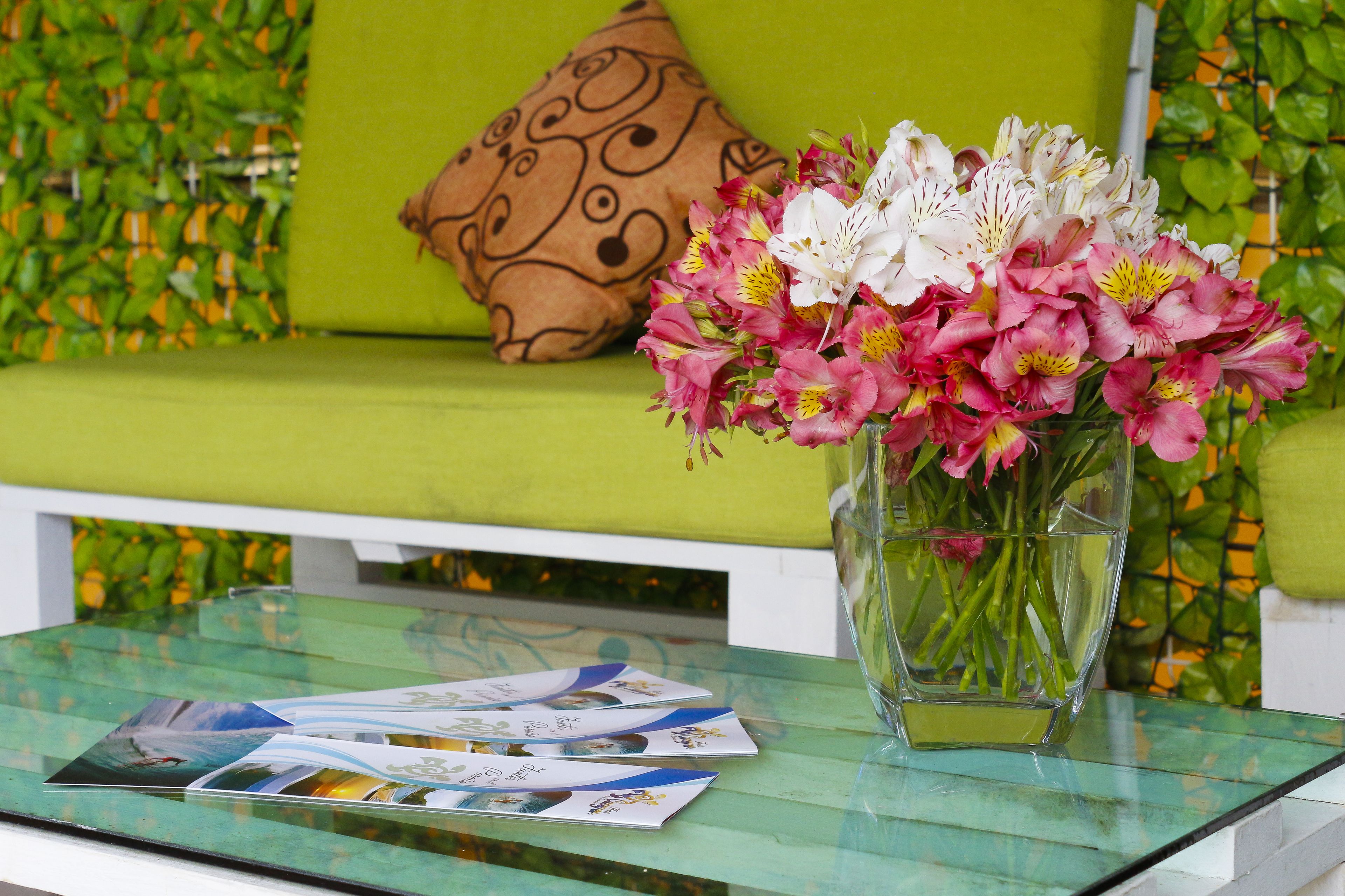 Een glazen tafel met een vaas roze en witte bloemen, een gestreept kussen en brochures op een groene, beklede bank.