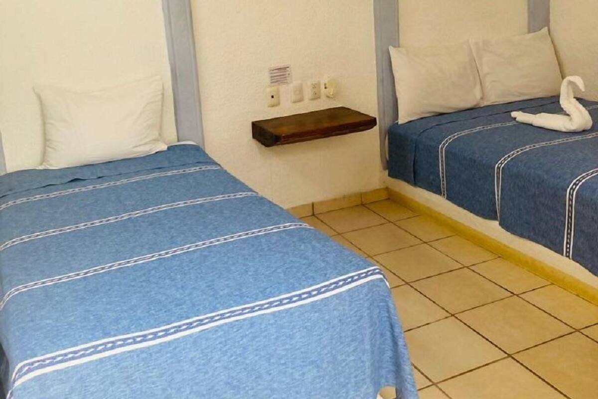 Une chambre avec deux lits, chacun recouvert d’une courtepointe bleue et agrémenté d’oreillers blancs. On y trouve une petite étagère en bois où sont posés des articles de toilette.