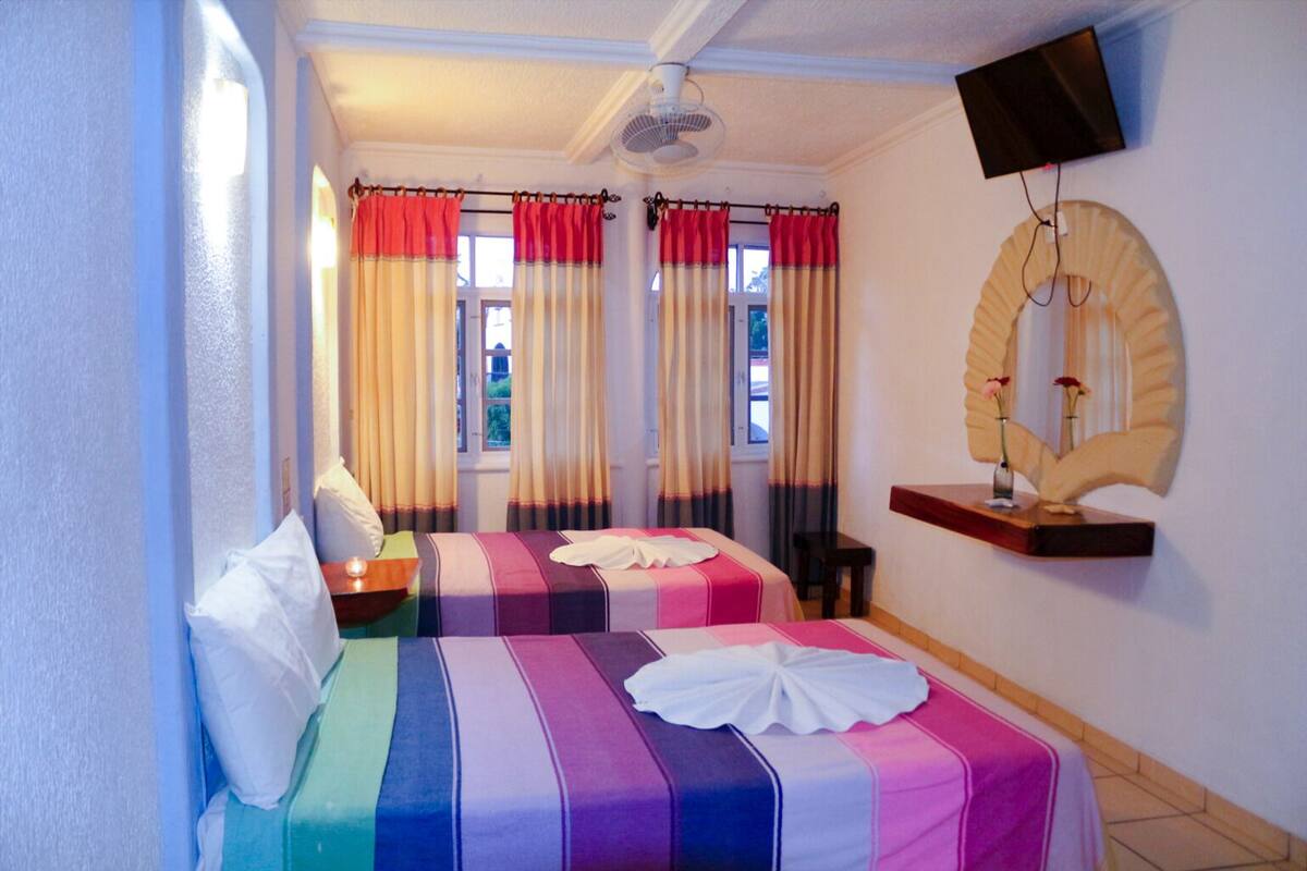 Une chambre d’hôtel avec deux lits, un ventilateur de plafond, une télévision et un miroir agrémenté d’un vase de fleurs.