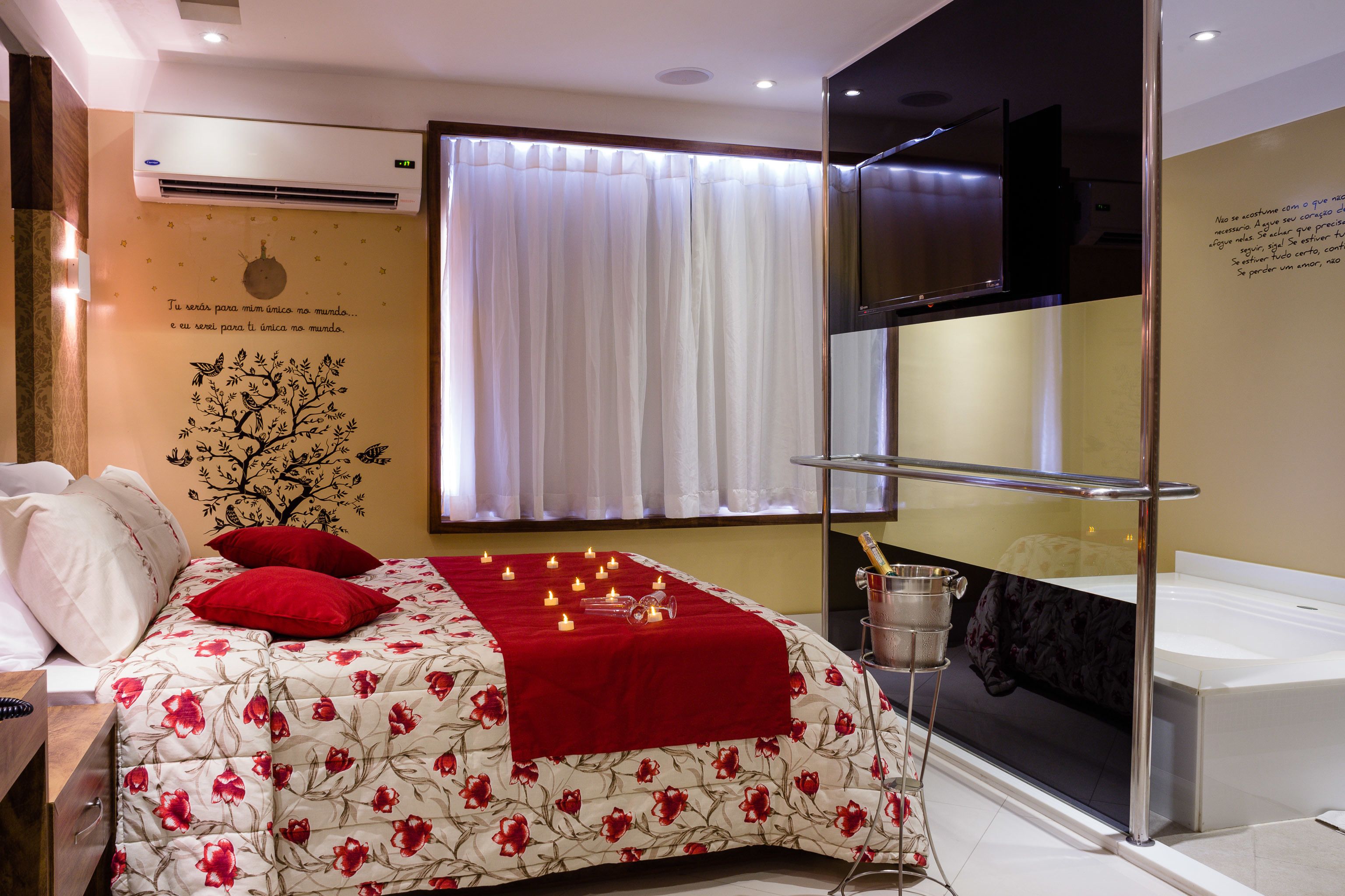 A hotel room with a bed, red pillows, a red blanket, a wall with a tree decal, a window with white curtains, a TV, and a minibar.
