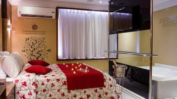 Habitación de hotel con cama, almohadas y manta rojas, pared decorada con un adhesivo de árbol, ventana con cortinas blancas, televisor y minibar.