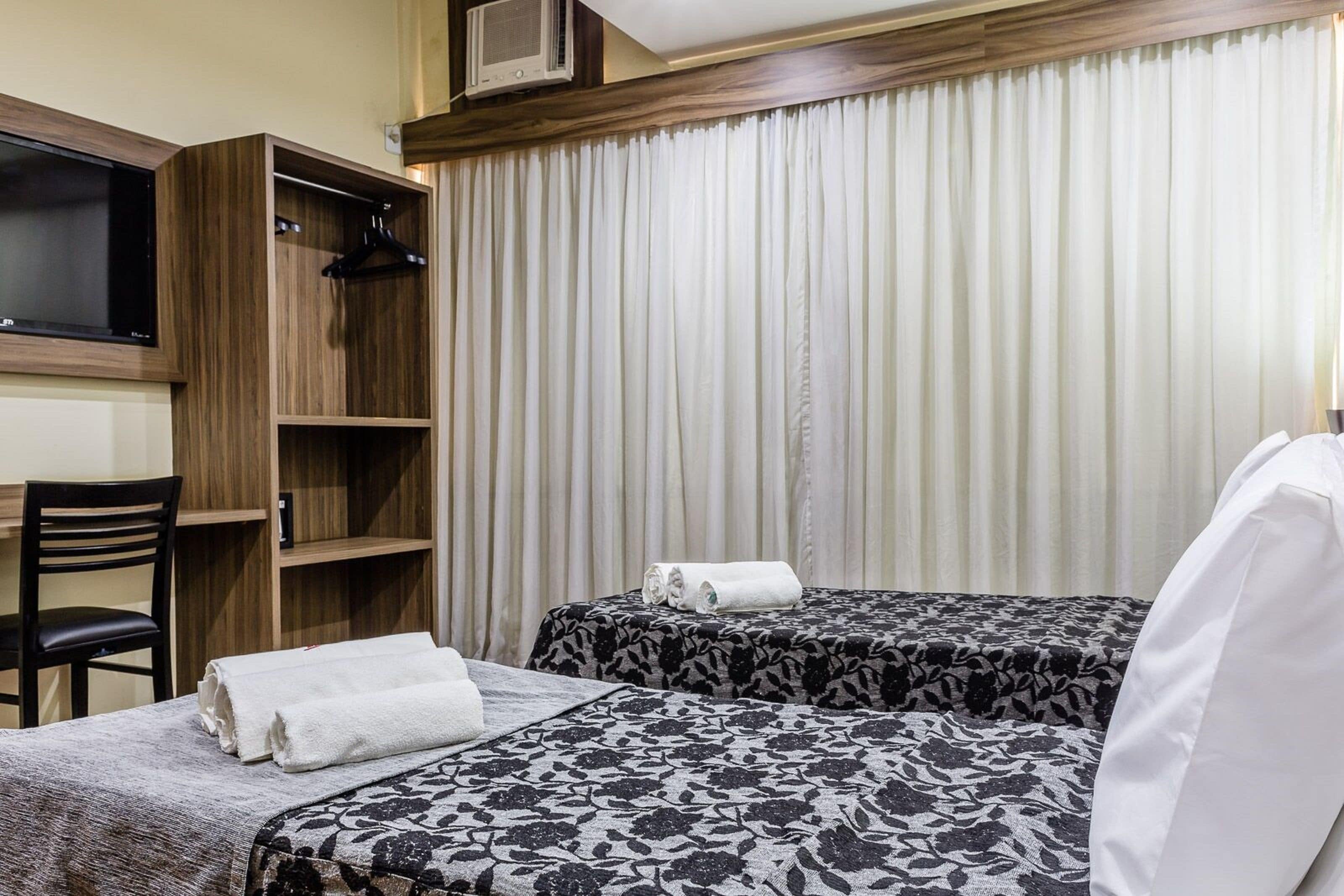 Une chambre d’hôtel avec deux lits, une télévision, une armoire en bois, une chaise et une grande fenêtre avec des rideaux.