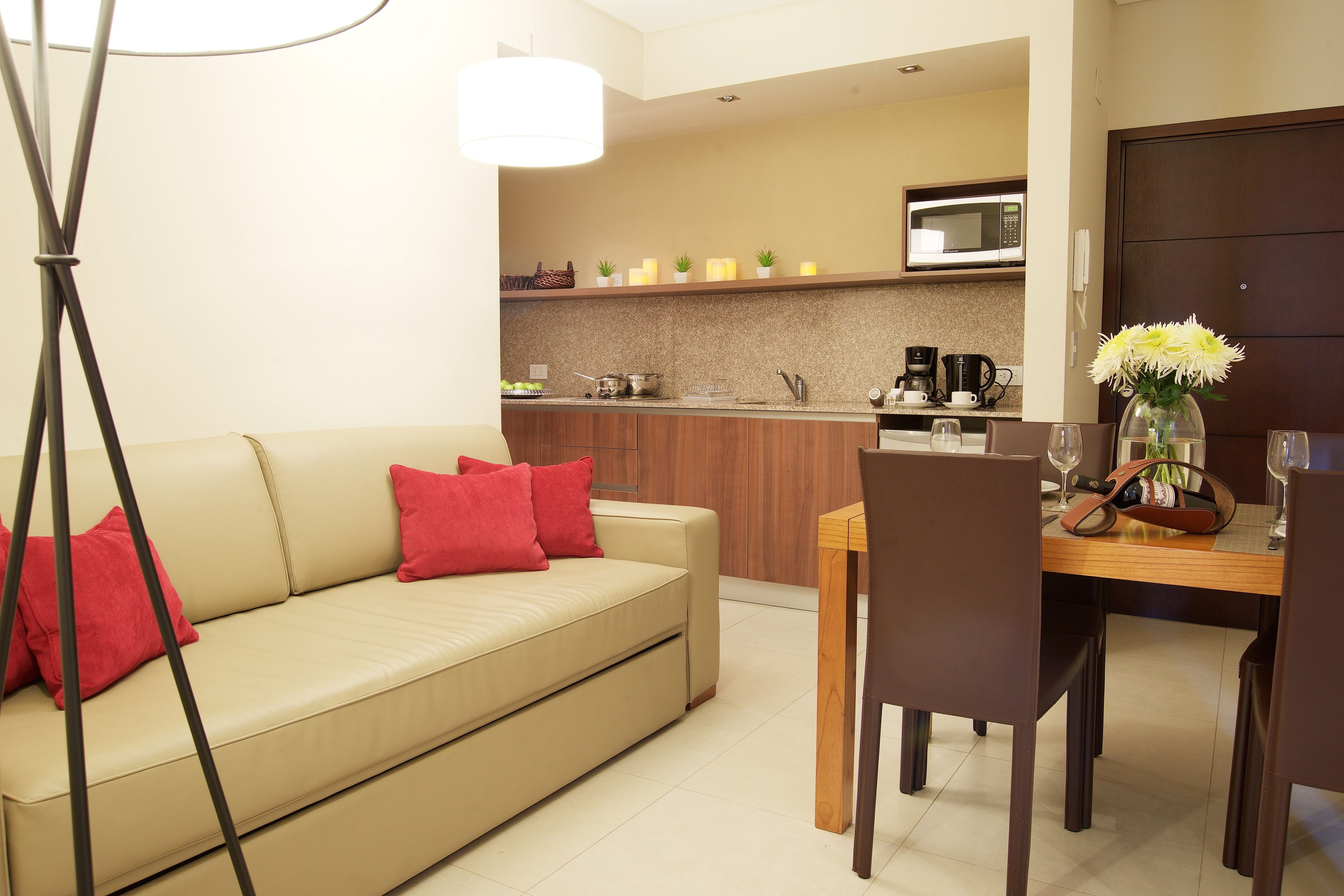Een moderne woonkamer met een beige bank, rode kussens, een eettafel met stoelen, een keuken met een magnetron, en een vaas met bloemen.