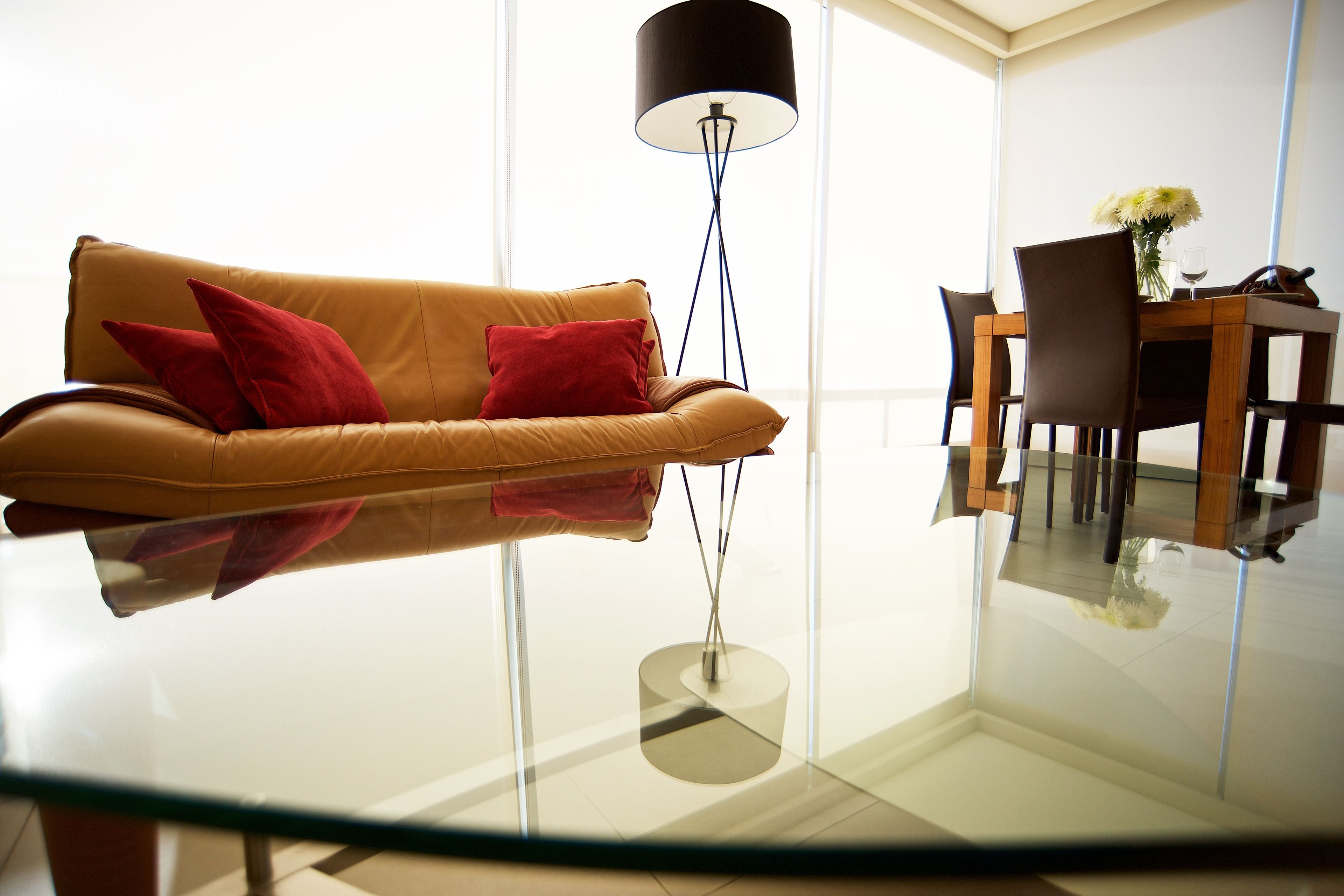 Una sala moderna con una mesa de centro de vidrio, un sofá de cuero marrón con cojines rojos, una lámpara de pie negra y un comedor con una mesa y sillas de madera.