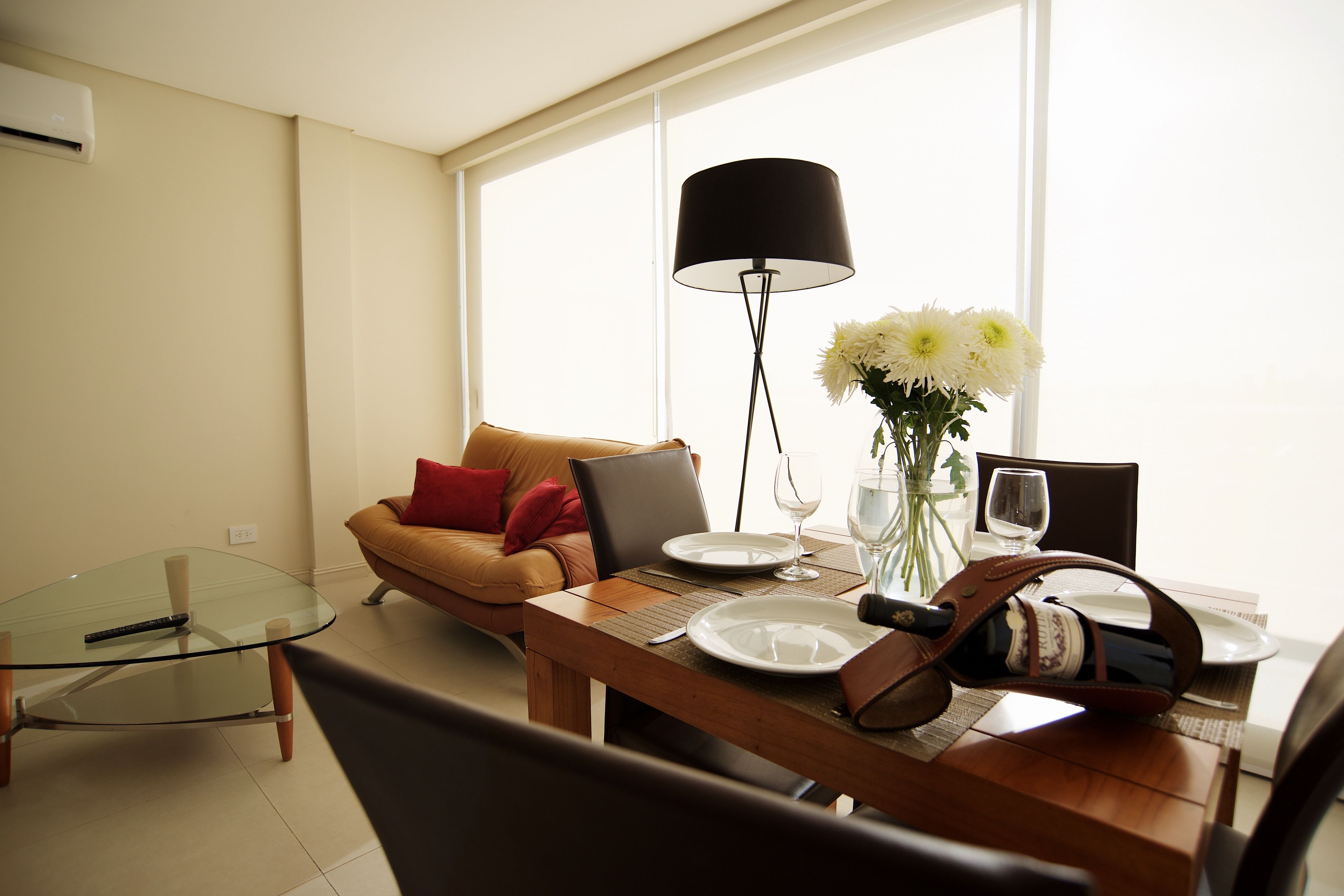 Een moderne woonkamer met een glazen salontafel, een bruine leren bank met rode kussens, een eettafel gedekt met borden, glazen en een laptop.