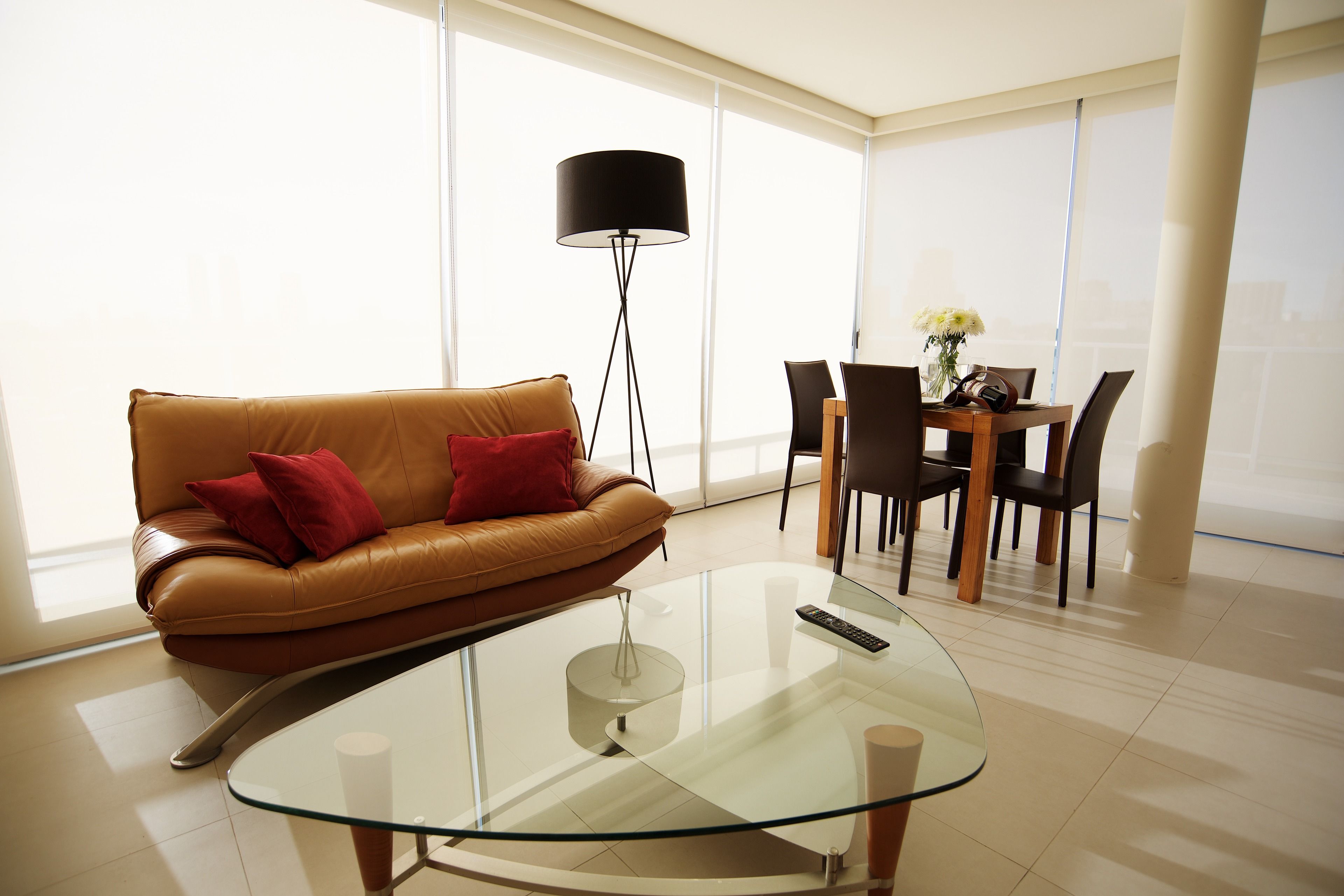 A modern living room with a brown leather sofa, a glass coffee table, a black floor lamp, and a dining area with a wooden table and chairs.