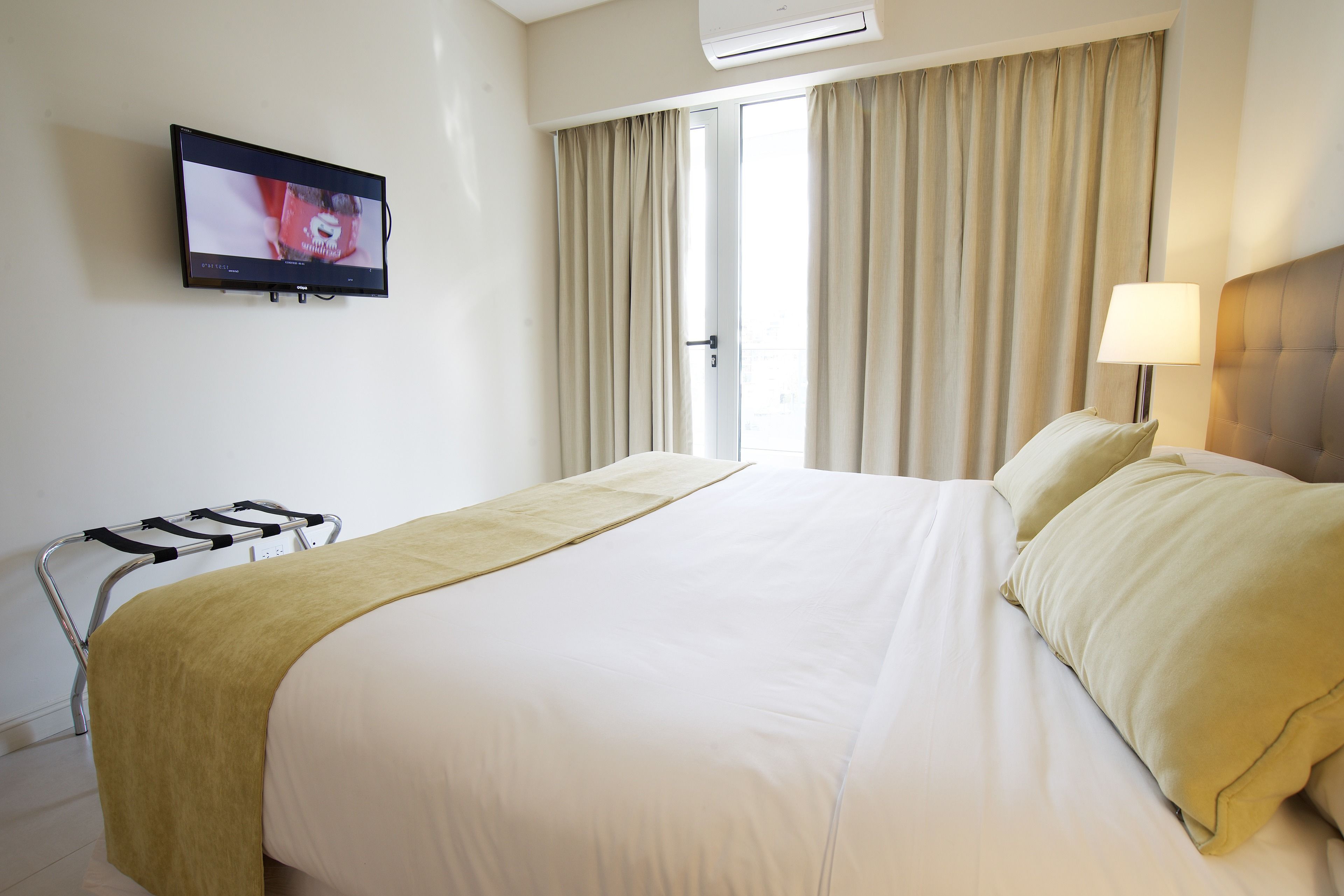 Habitación de hotel con cama, televisor montado en la pared, ventana con cortinas y lámpara de noche.
