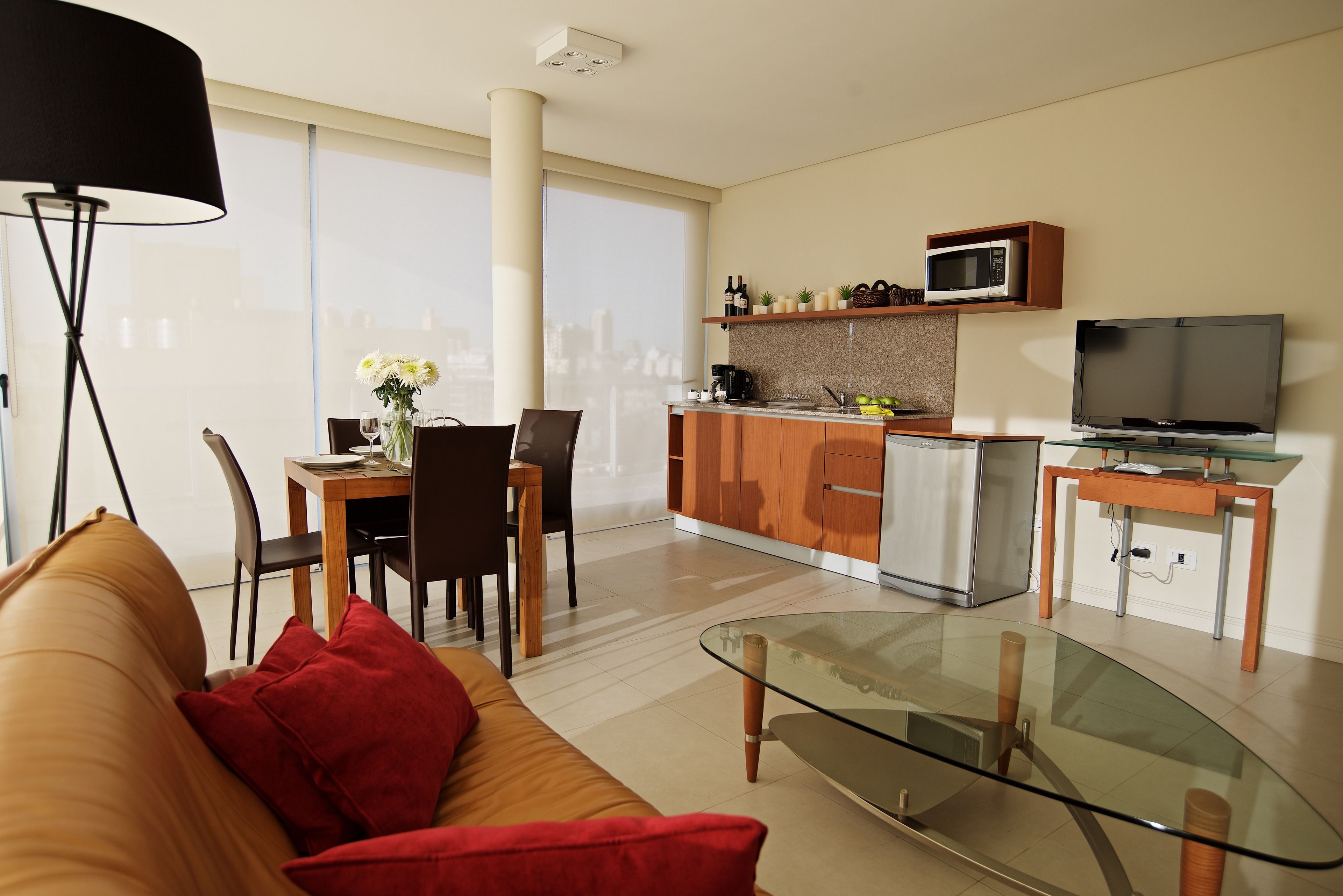 A modern living room with a glass coffee table, a sofa with red cushions, a dining area with a table and chairs, a kitchen with a microwave and a TV, and large windows offering a city view.