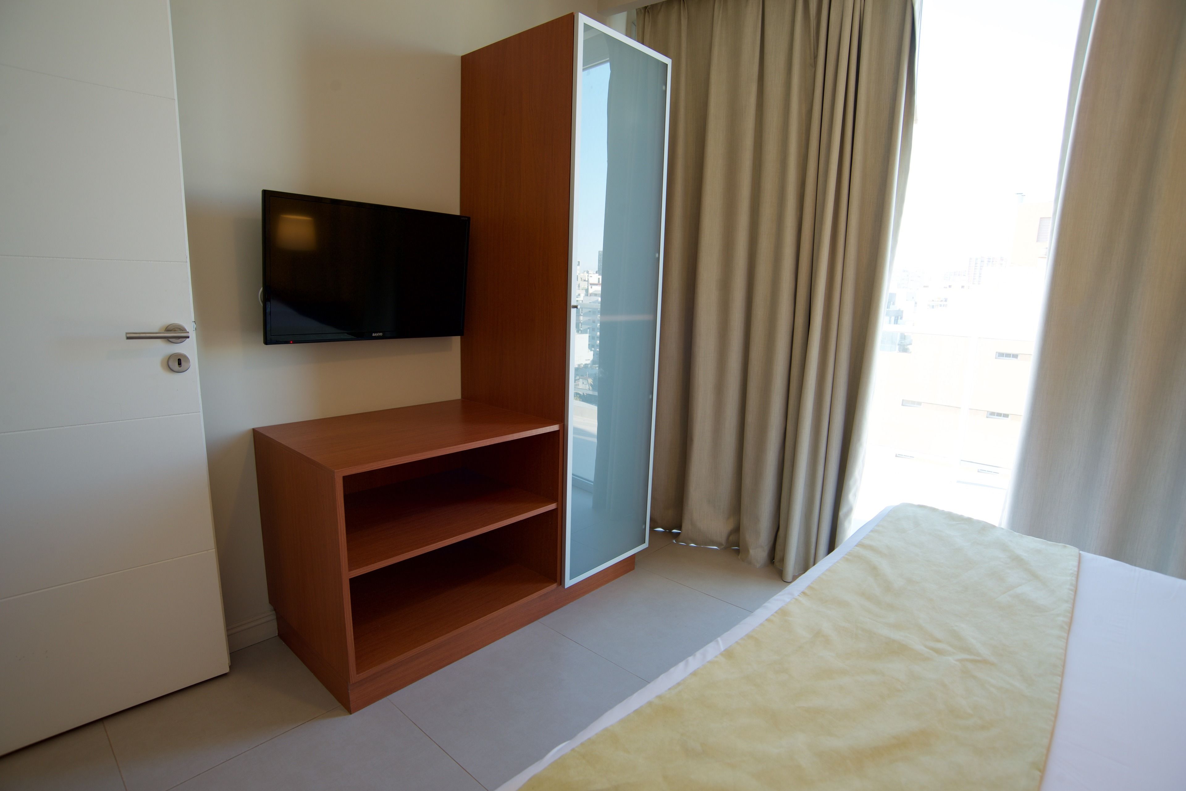 A hotel room with a bed, a wooden cabinet with shelves, a television on the wall, and a window with curtains.