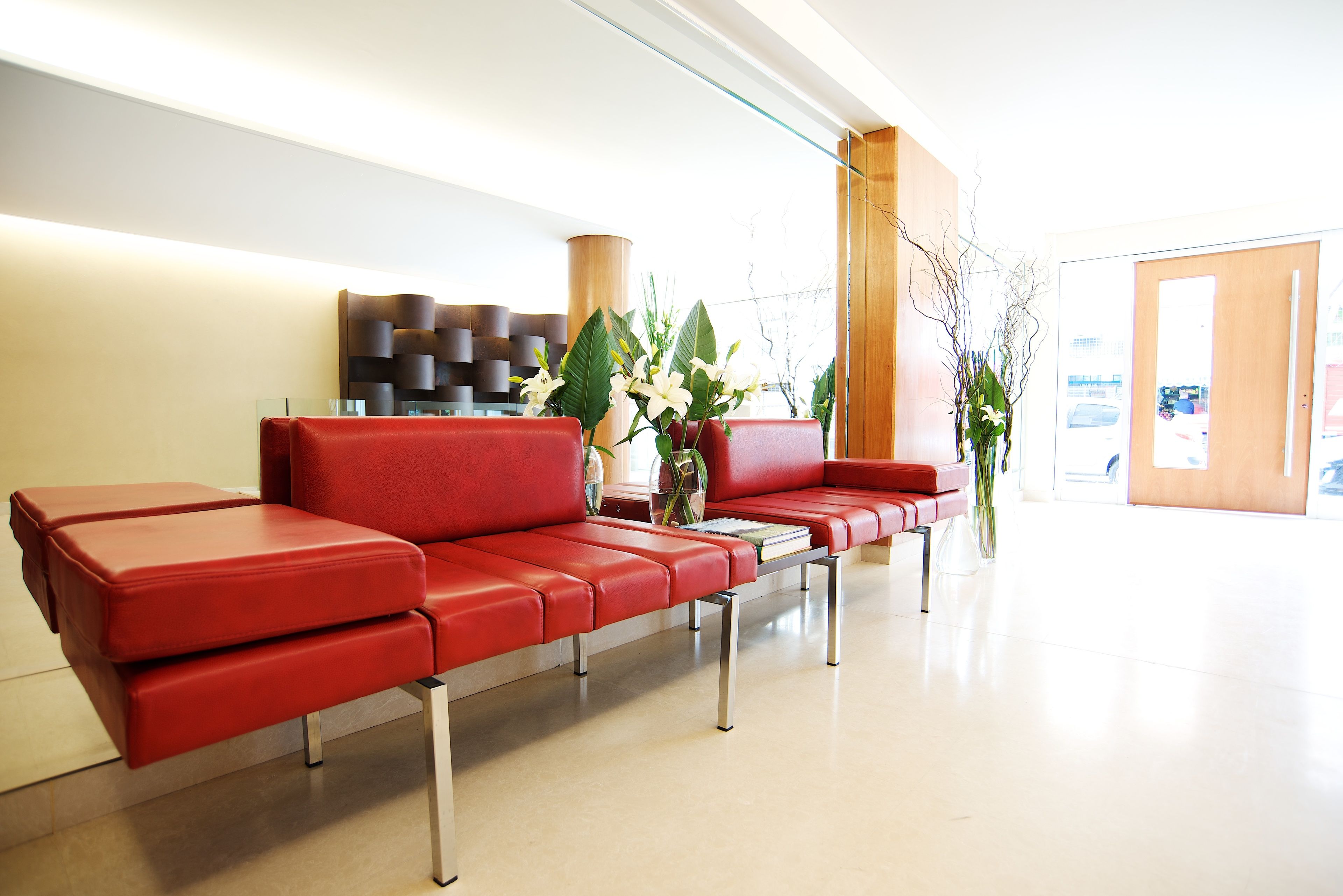 Un espace d’attente moderne avec des sièges en cuir rouge, une table basse en verre et des plantes décoratives.