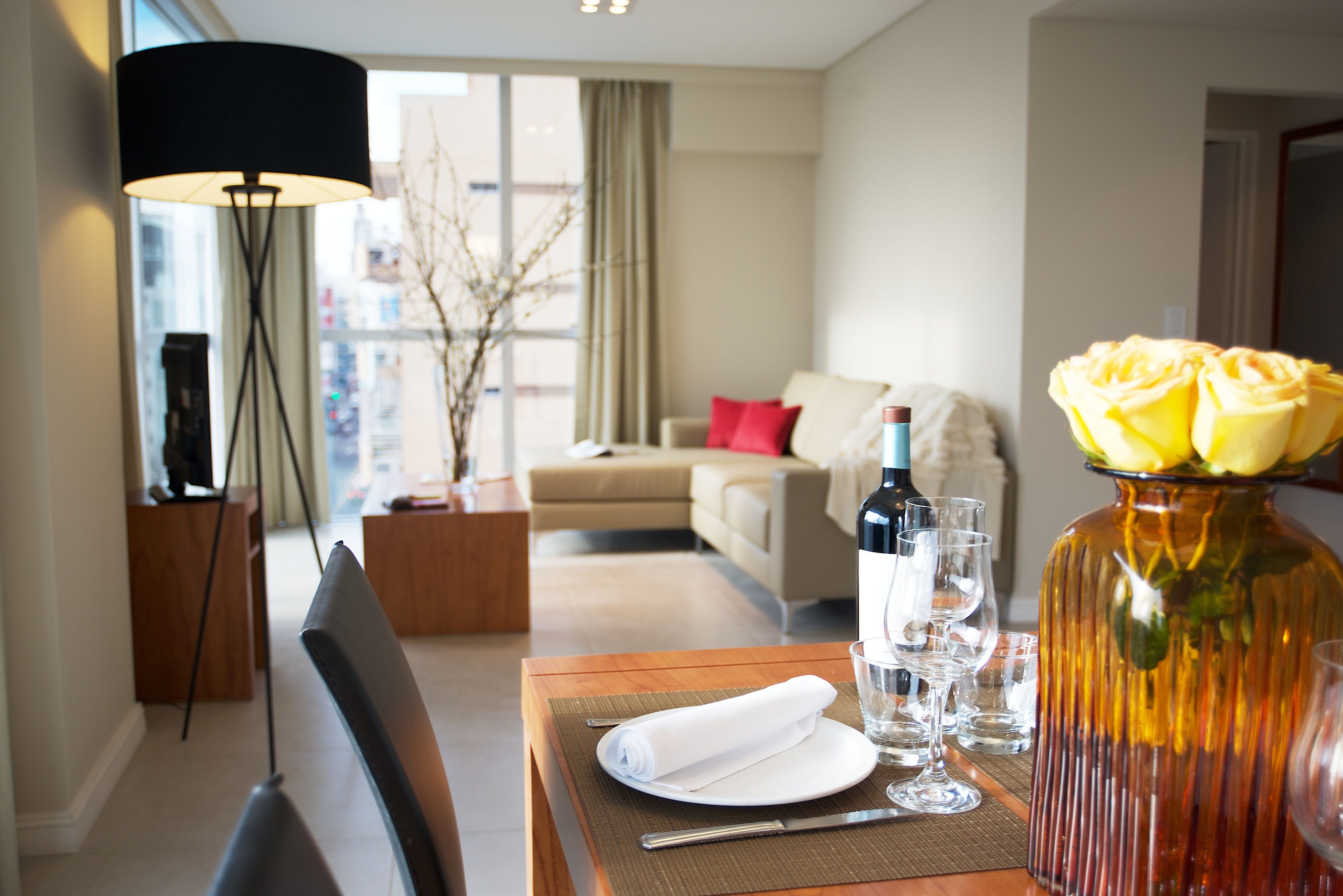 A hotel room with a dining table set for two, a sofa, a lamp, and a vase with yellow flowers.