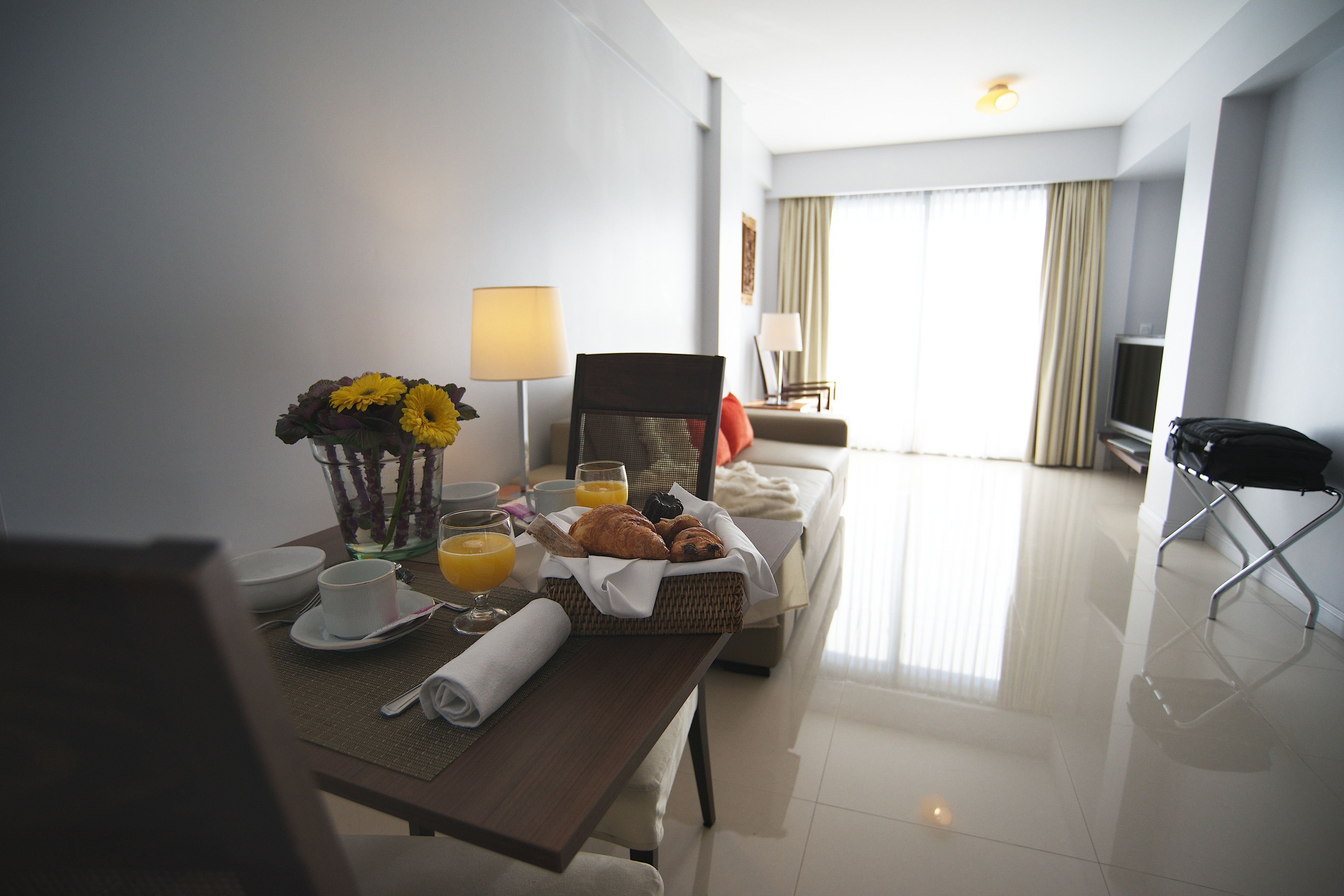A hotel room with a dining table set with breakfast items, a TV, and a bed with a view of the city.