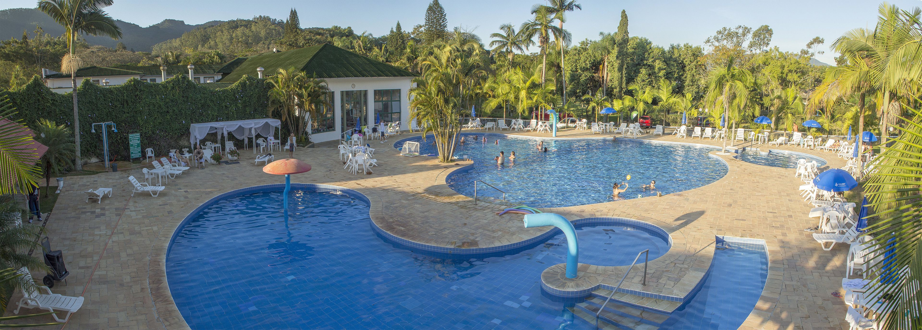 Una amplia piscina al aire libre con múltiples carriles, rodeada de sillones de descanso y sombrillas, con palmeras y un paisaje montañoso de fondo.