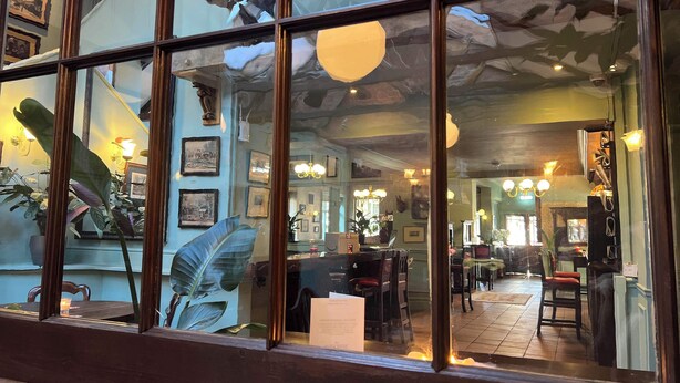 Interior de restaurant amb una gran finestra de vidre, mobles de fusta i diverses imatges emmarcades a les parets.