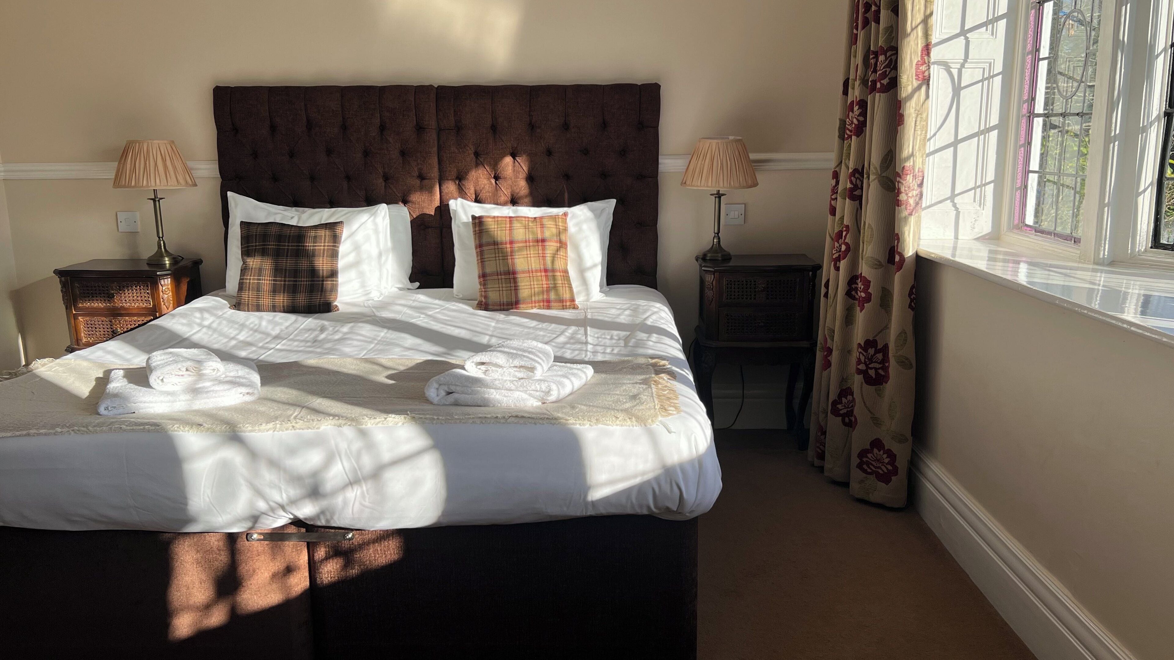Una cama bien tendida con sábanas blancas y una toalla doblada, flanqueada por dos mesitas de noche con lámparas, en una habitación con un gran ventanal y cortinas florales.