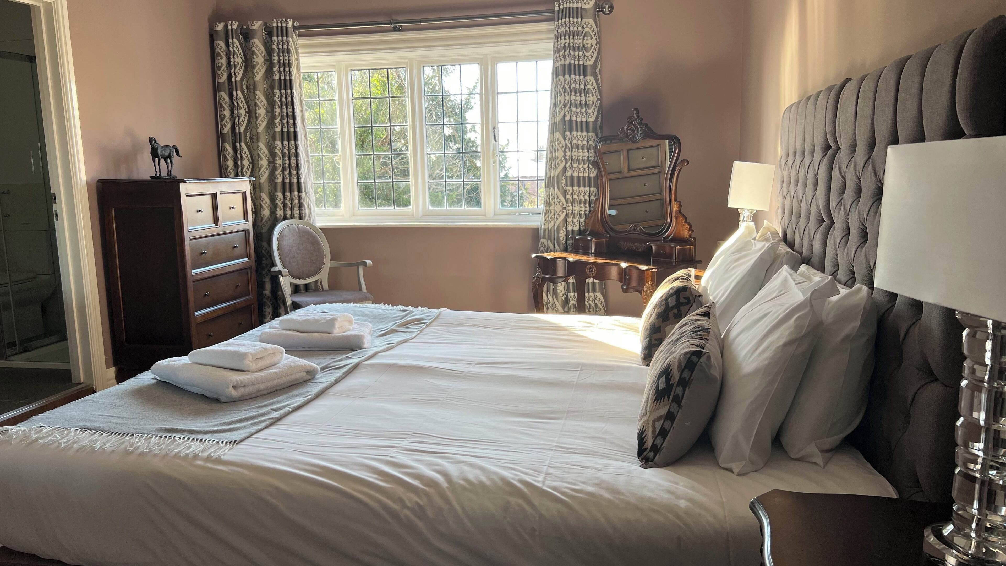A neatly made bed with white linens and towels, a wooden dresser, a chair, and a desk with a lamp.