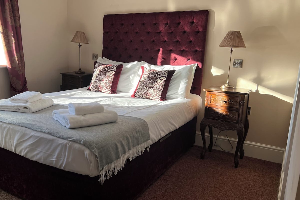 Una cama bien hecha con sábanas blancas y un plaid gris, un cabecero rojo oscuro y almohadones decorativos. Hay dos mesitas de noche con lámparas, y una ventana con cortinas.