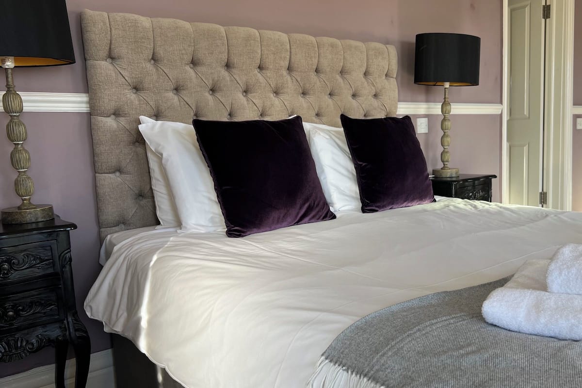 Una cama bien hecha con sábanas blancas y dos almohadas moradas oscuras, una mesita de noche con una lámpara y un radiador al fondo.