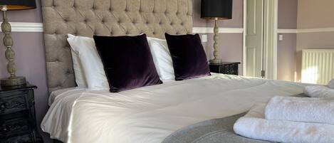 A neatly made bed with white linens and two dark purple pillows, a bedside table with a lamp, and a radiator in the background.
