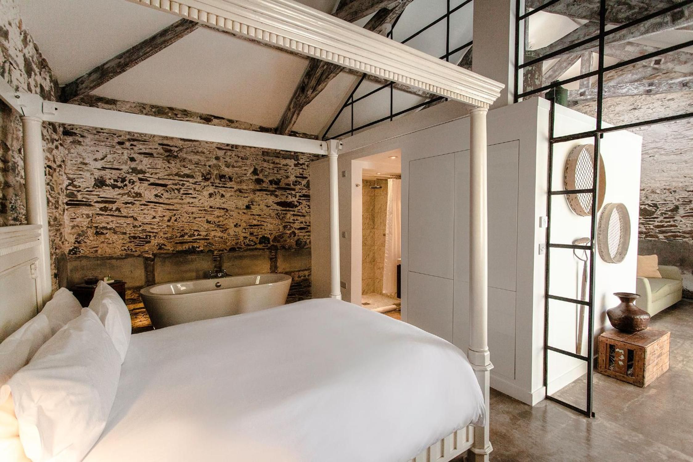 A bedroom with a stone wall, a large bed, a bathtub, and a view through an open door.