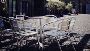 Ein Sitzbereich im Freien mit Metallstühlen und -tischen, einer Holzw pergola und Gebäuden aus Stein im Hintergrund.