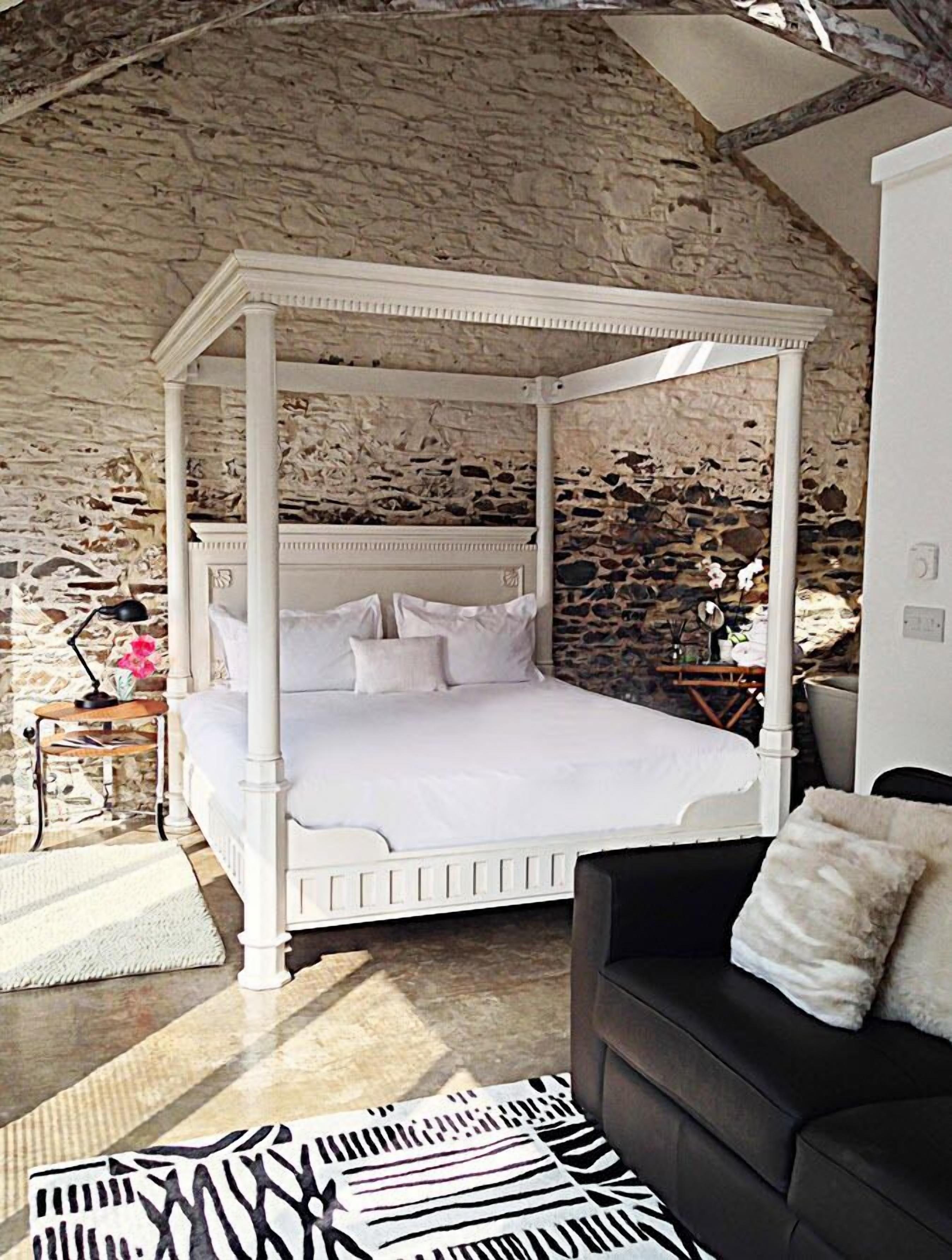 Un dormitorio con cama con dosel, paredes de piedra, una mesita con lámpara y una alfombra con estampado de cebra.