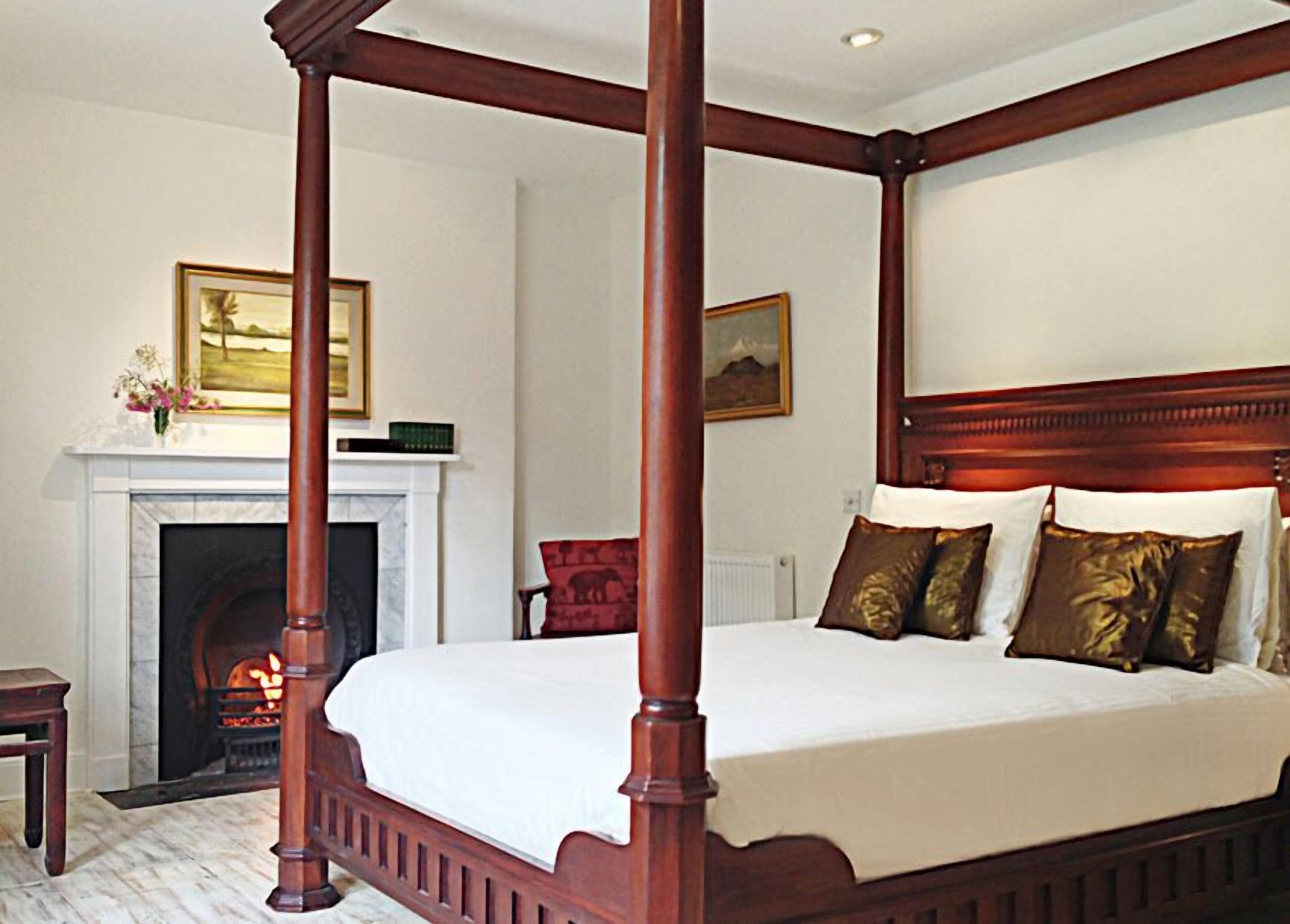 Una cama con dosel con cubrecama blanco y almohadas marrones, una chimenea con repisa, y un cuadro de un paisaje colgado sobre ella.