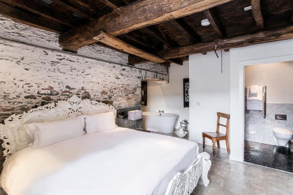 A bedroom with a stone wall, a bed with ornate headboard, a bedside table, a chair, and a bathroom with a sink and toilet.