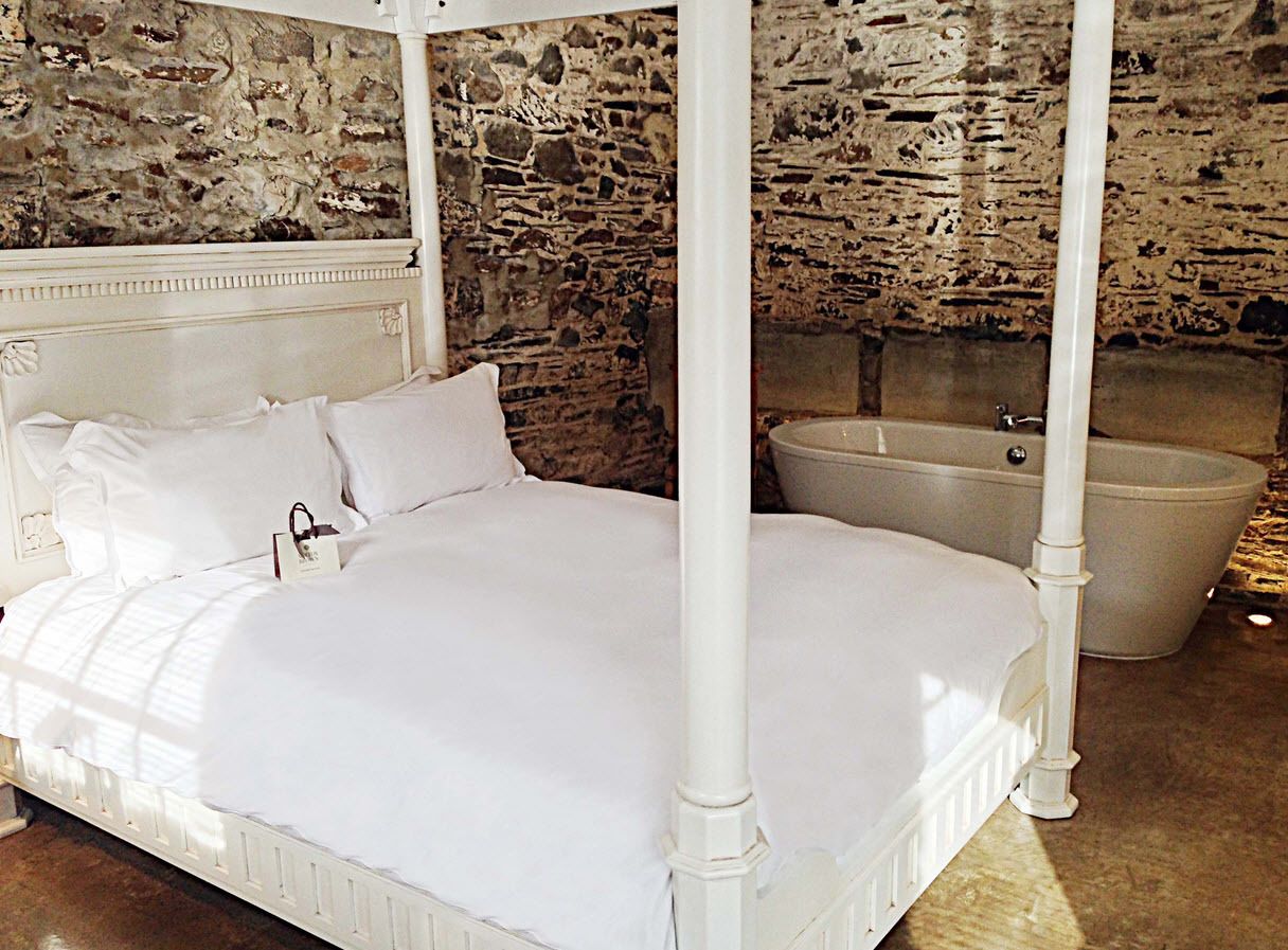 A four-poster bed with white bedding in a room with stone walls and a bathtub.