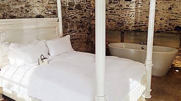 A four-poster bed with white bedding in a room with stone walls and a bathtub.