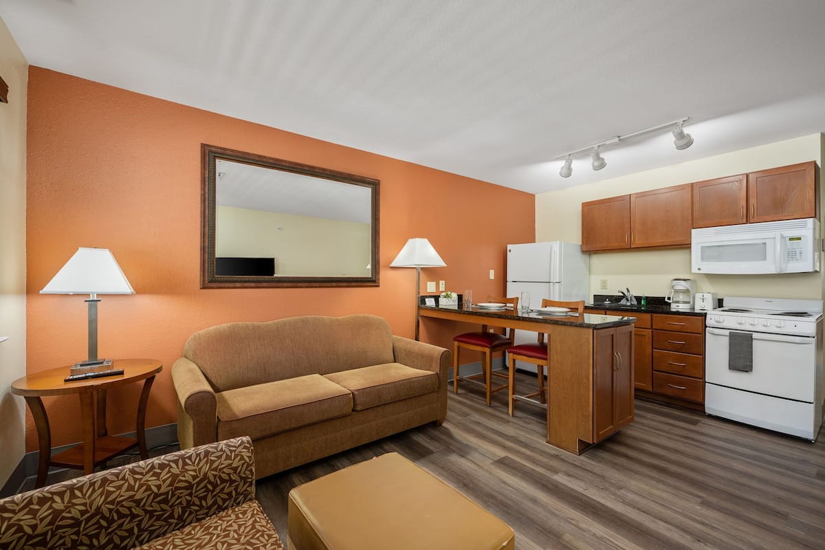 Una habitación de hotel compacta con kitchenette, sofá, mesa de comedor y espejo.