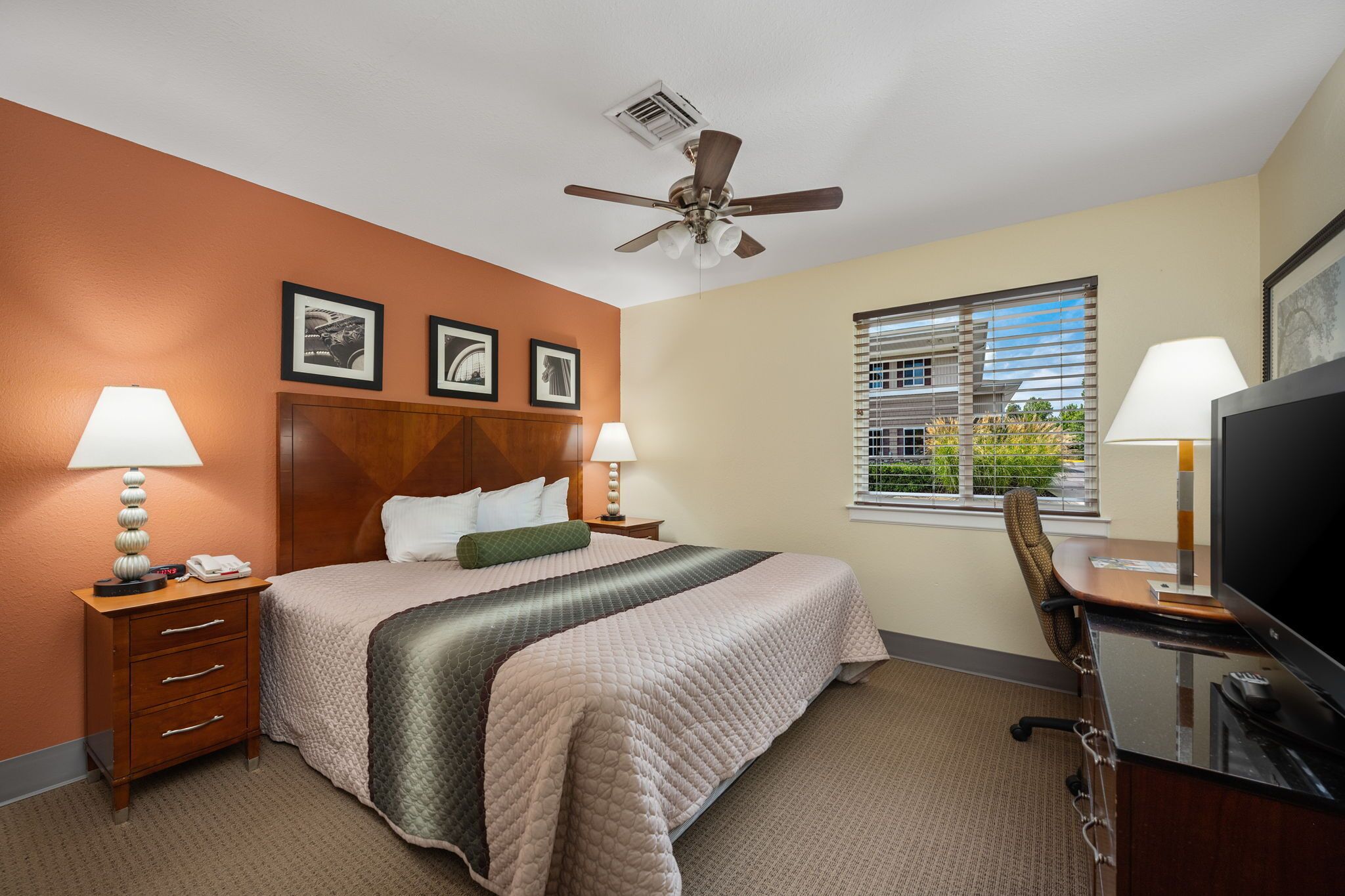 A hotel room with a bed, two bedside tables with lamps, a desk with a TV, a ceiling fan, and a window with blinds.