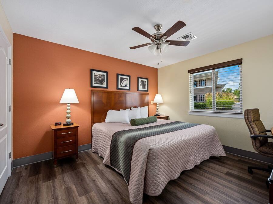 Quarto de hotel com uma cama, duas mesas de cabeceira com luminárias, um ventilador de teto, uma janela com persianas e vista para uma casa lá fora.