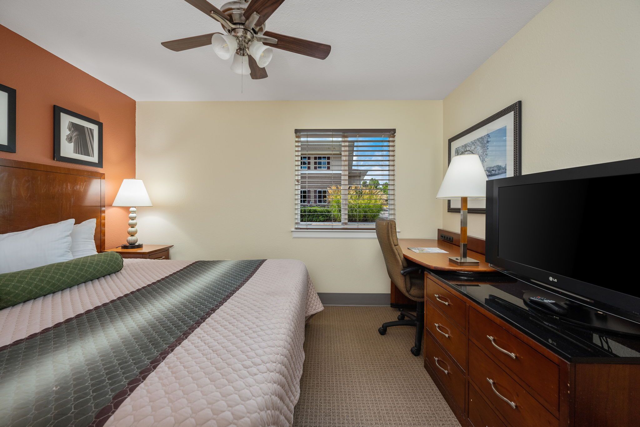 A hotel room with a bed, a desk with a TV, a chair, a window with blinds, and a ceiling fan.