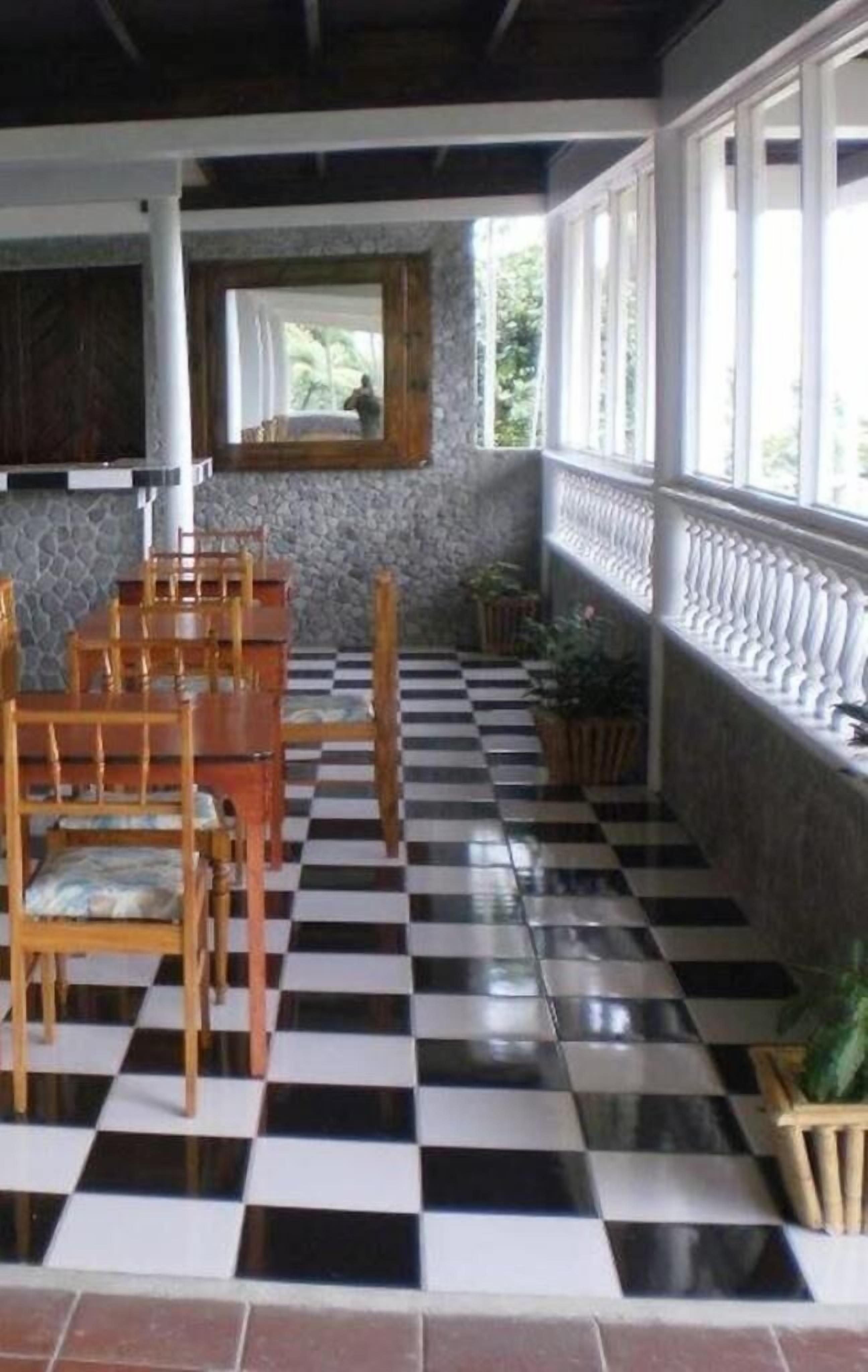 A room with a checkered floor, wooden chairs, a stone wall, and a window with a view of greenery.