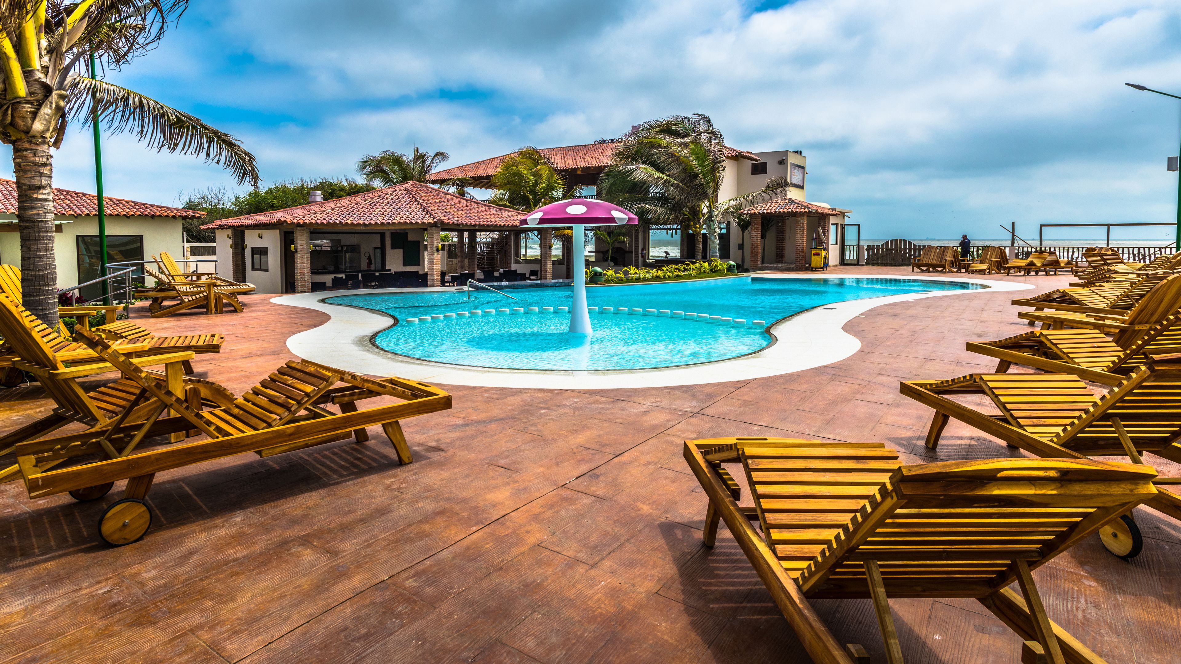 A pool area with wooden lounge chairs, a central water feature, and a building with a tiled roof in the background.