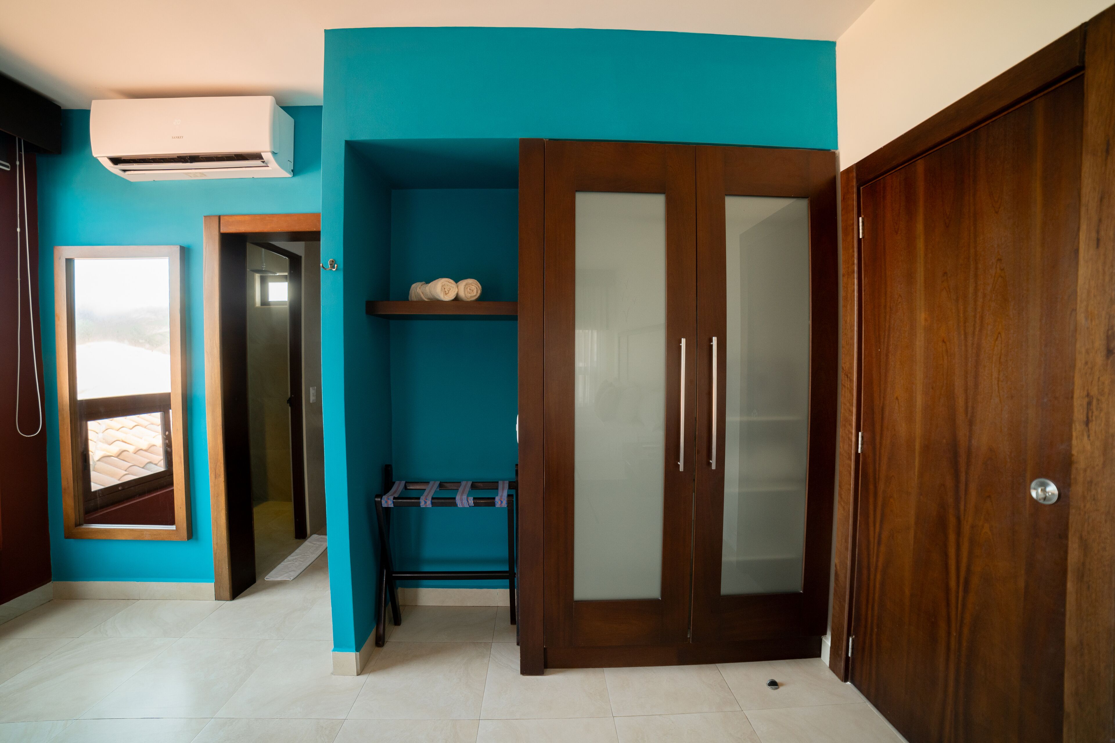 Ein Zimmer mit türkisfarbenen Wänden, einem Holzschrank, einer fest installierten Klimaanlage, einem Fenster und einer Tür, die in ein weiteres Zimmer führt.